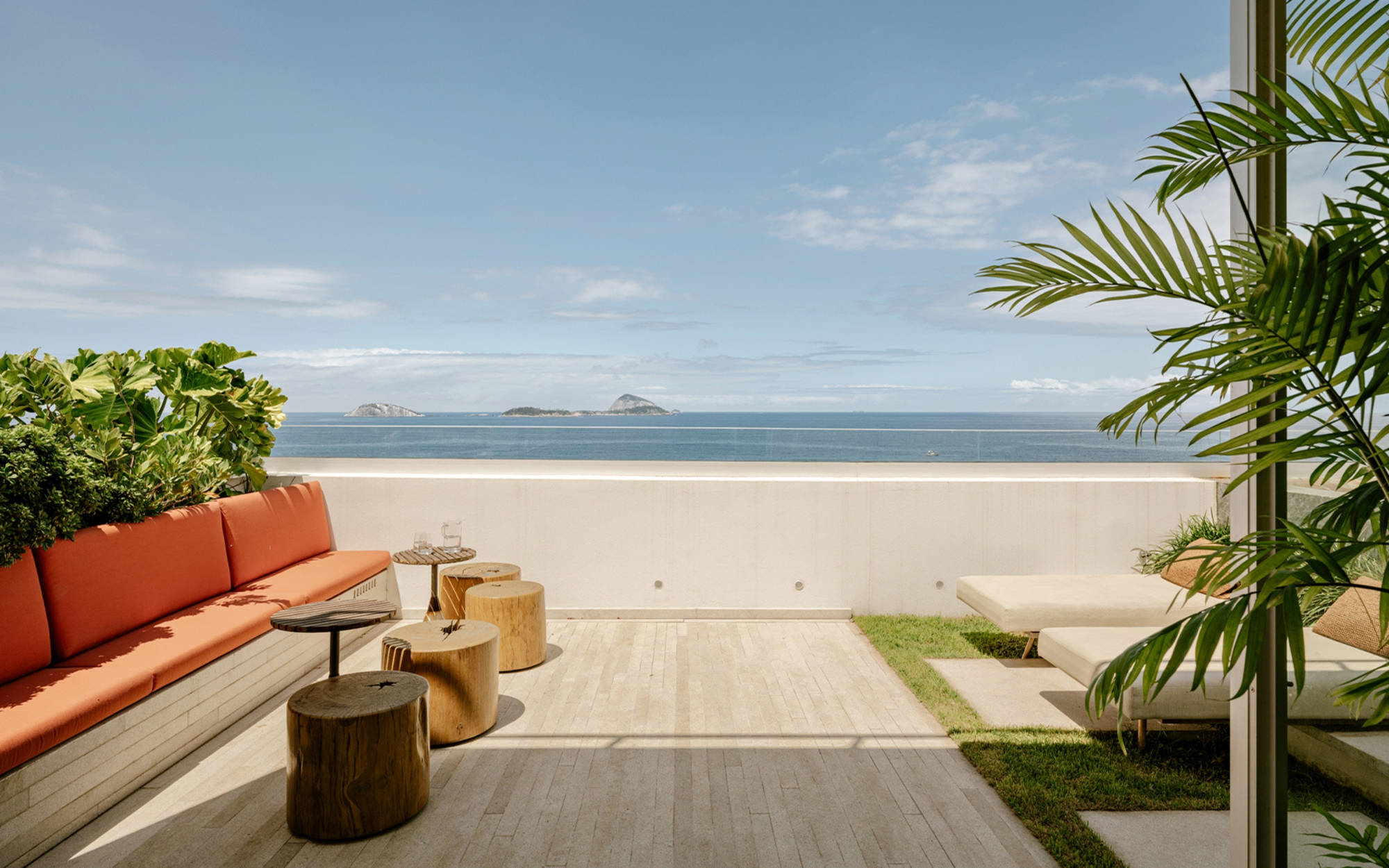 Terrace with a built-in orange cushioned bench, log stools and loungers on grass, facing the sea and distant islands under blue sky