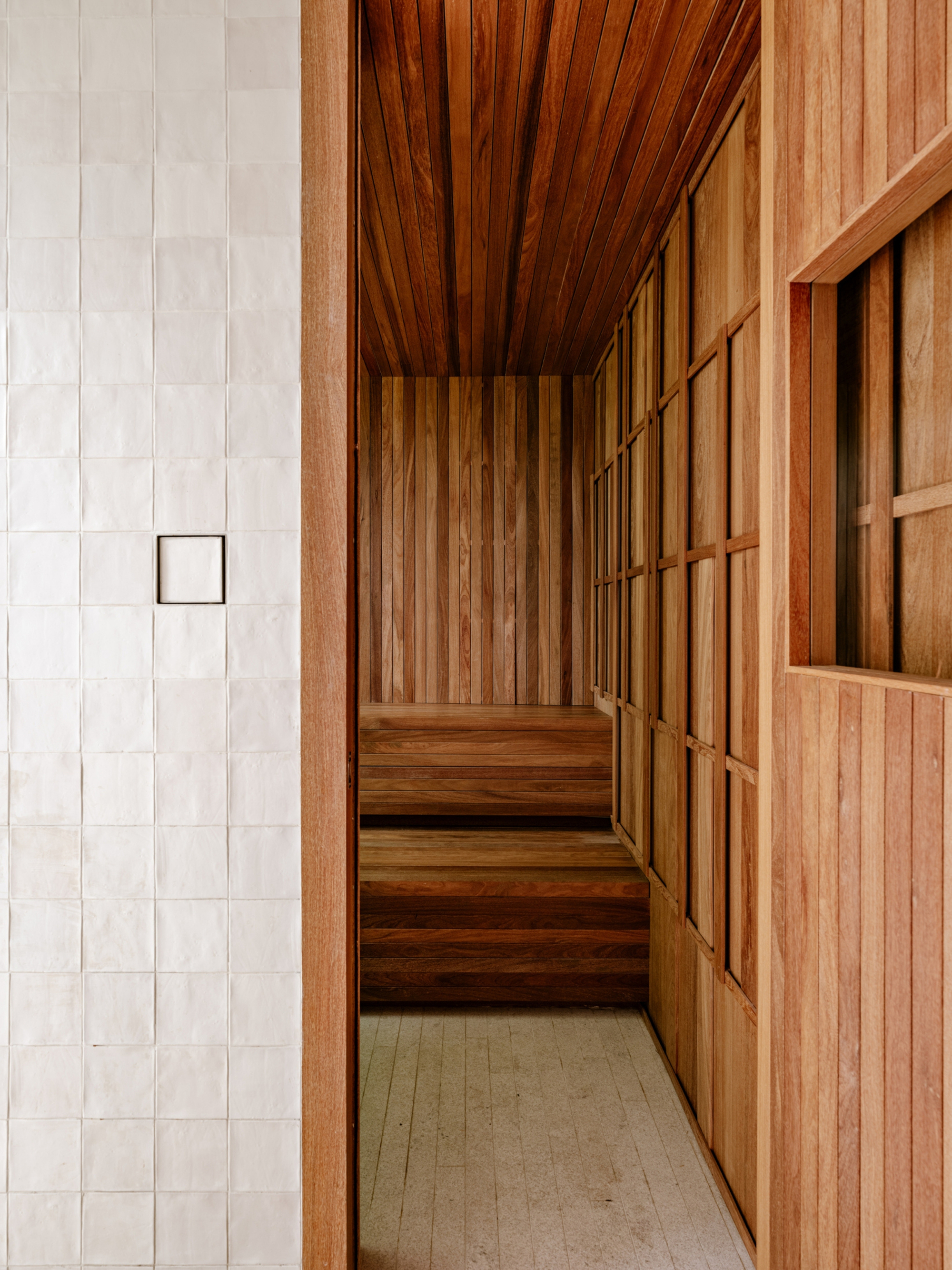 Timber-lined sauna with stepped benches seen past a glossy white tile wall, lit by soft side light