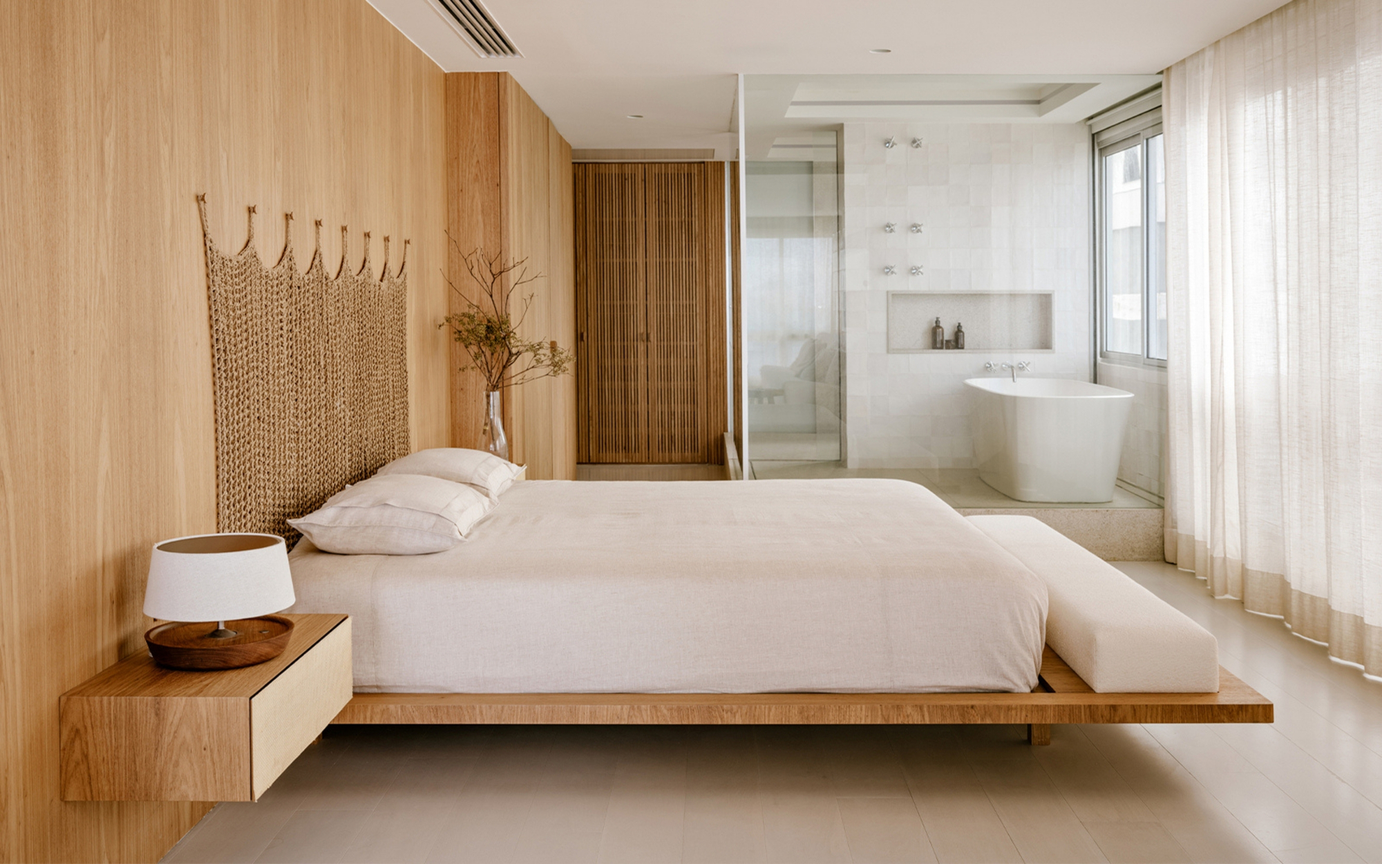 Low platform bed against an oak wall, with a freestanding tub behind glass and linen curtains at the window