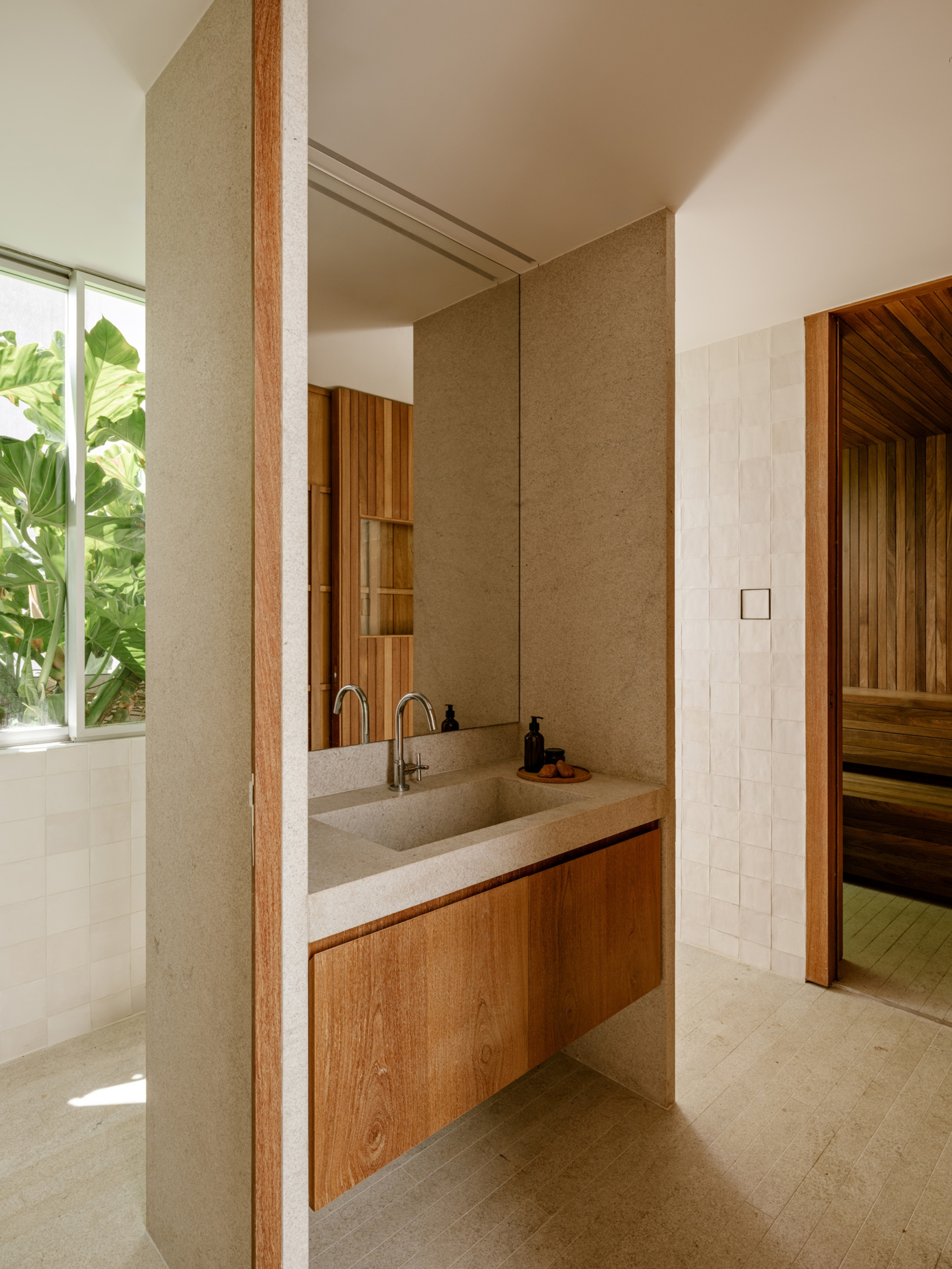 Stone basin with a floating timber drawer, mirrored above, beside a tiled wall and a window onto broadleaf plants