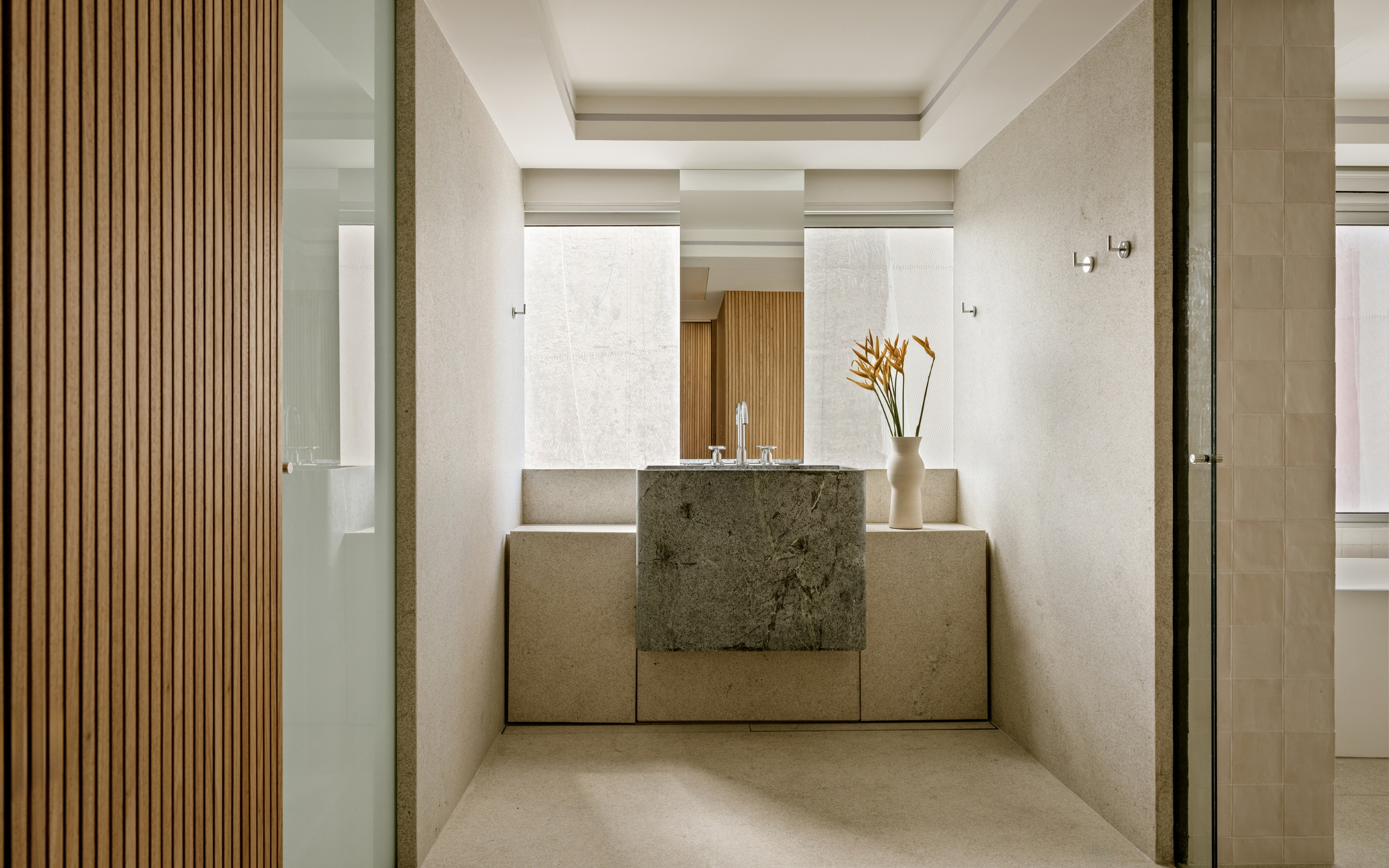 Bathroom vanity with a rough-cut green stone basin block, a heliconia stem in a white vase and a timber slat wall at left, lit by soft daylight