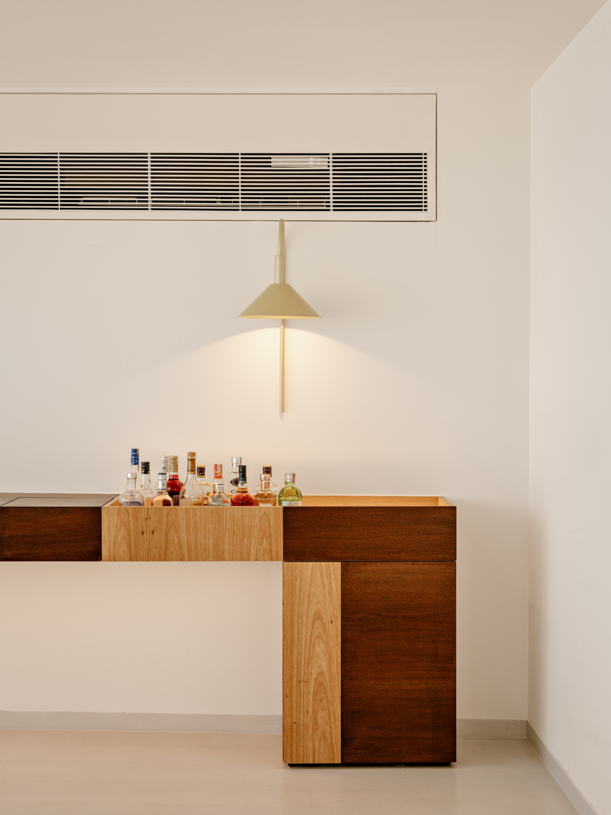 Wood bar console with two-tone timber panels and a row of liquor bottles, lit by a cone wall lamp beneath a ceiling vent