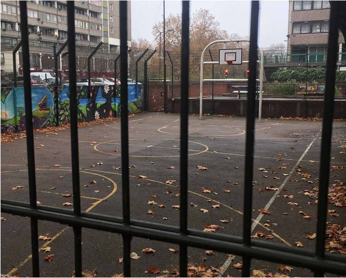 Existing asphalt basketball court seen through iron railings, with fallen autumn leaves, a mural wall and housing blocks behind. Image: Chris Moore