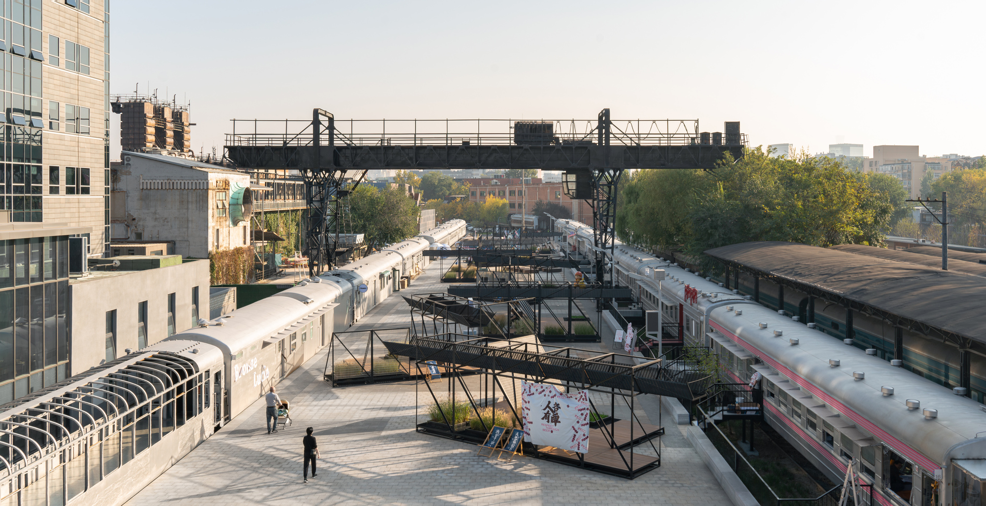 Elevated view of a plaza between old railway carriages, with a black steel gantry crane overhead and planted steel platforms in autumn light
