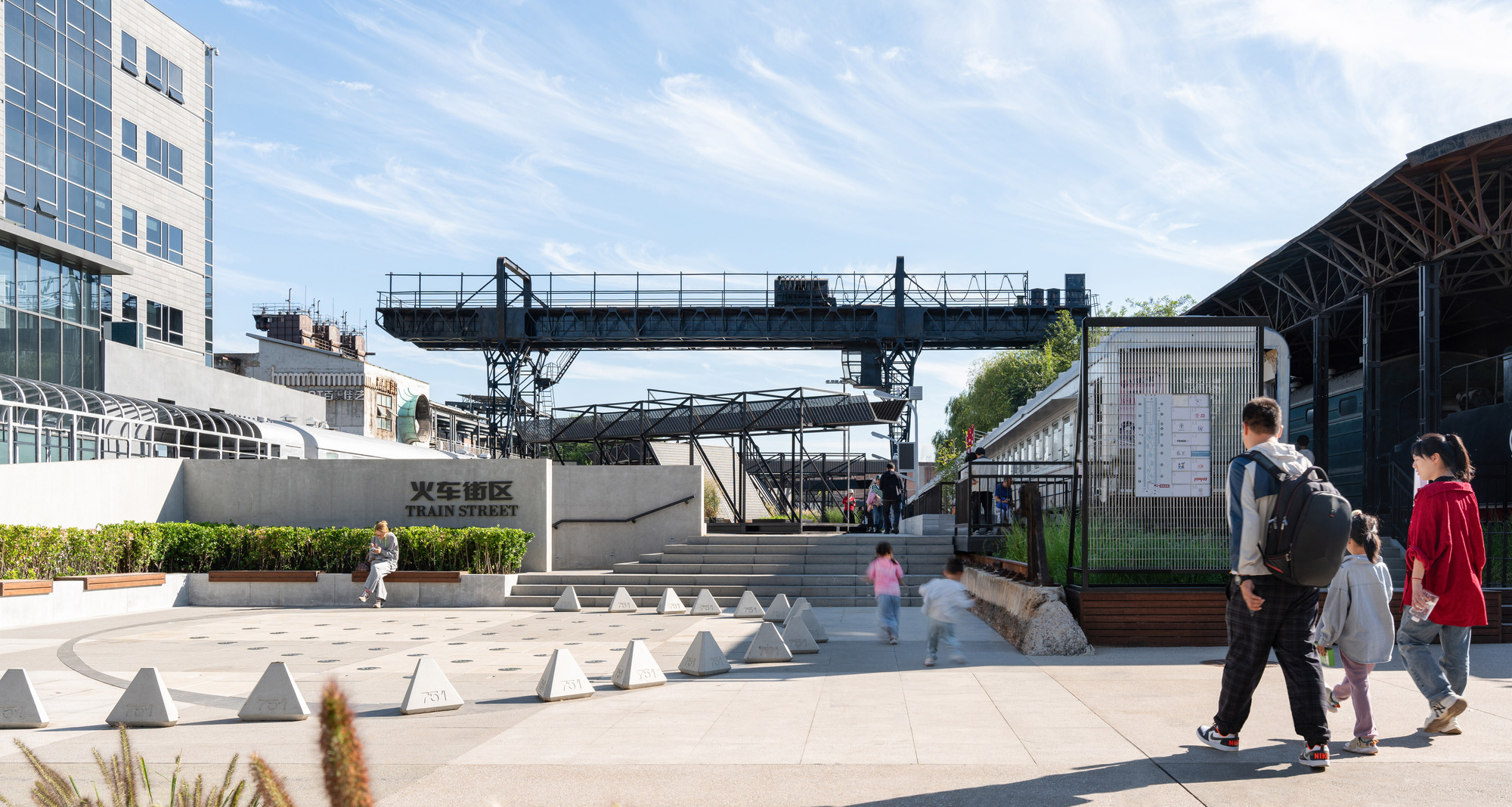 Entrance plaza with concrete bollards and a stepped ramp leading to the gantry crane, with families walking under a blue sky