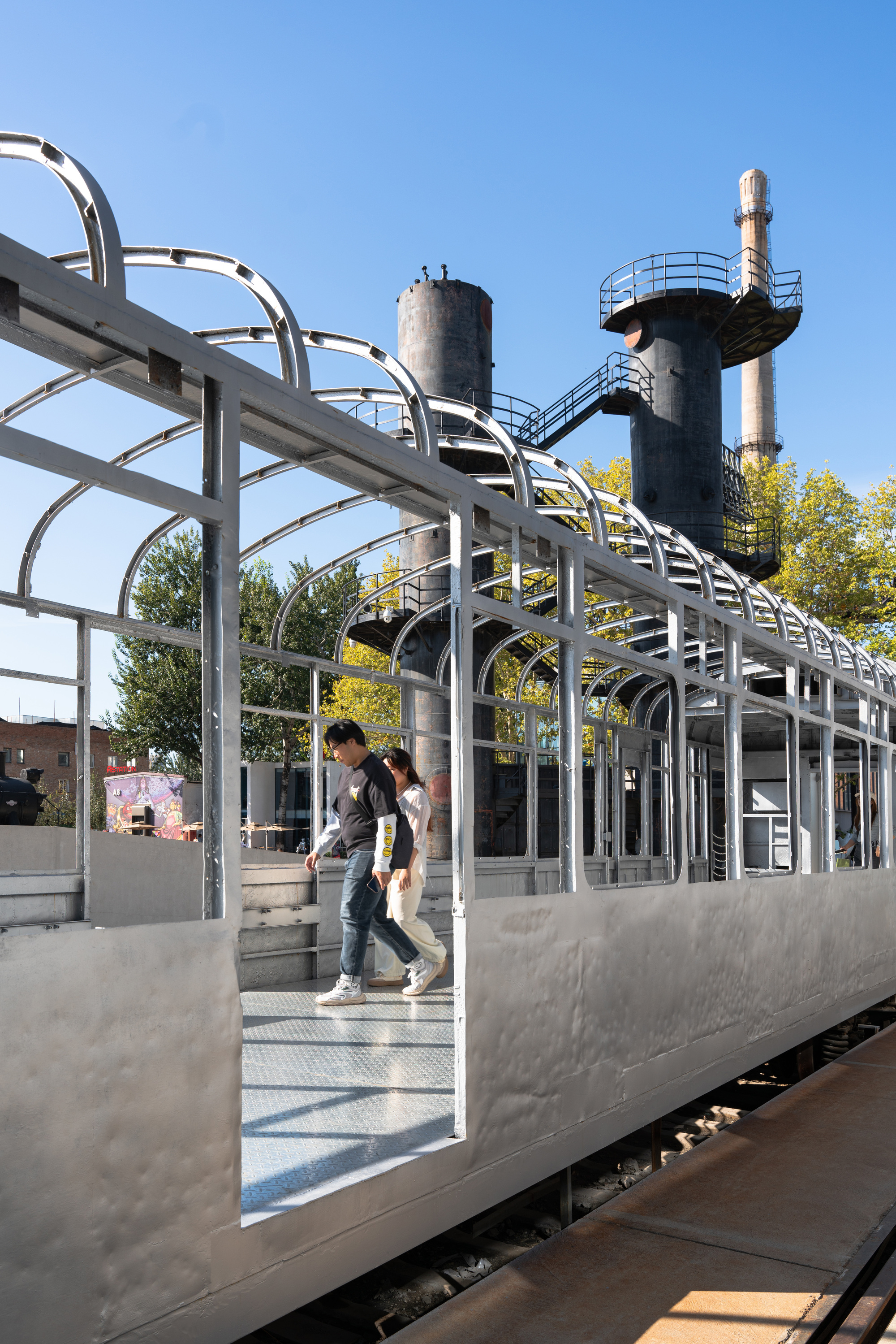 Two people step through the opening of a silver train carriage topped by steel arches, with dark storage tanks behind