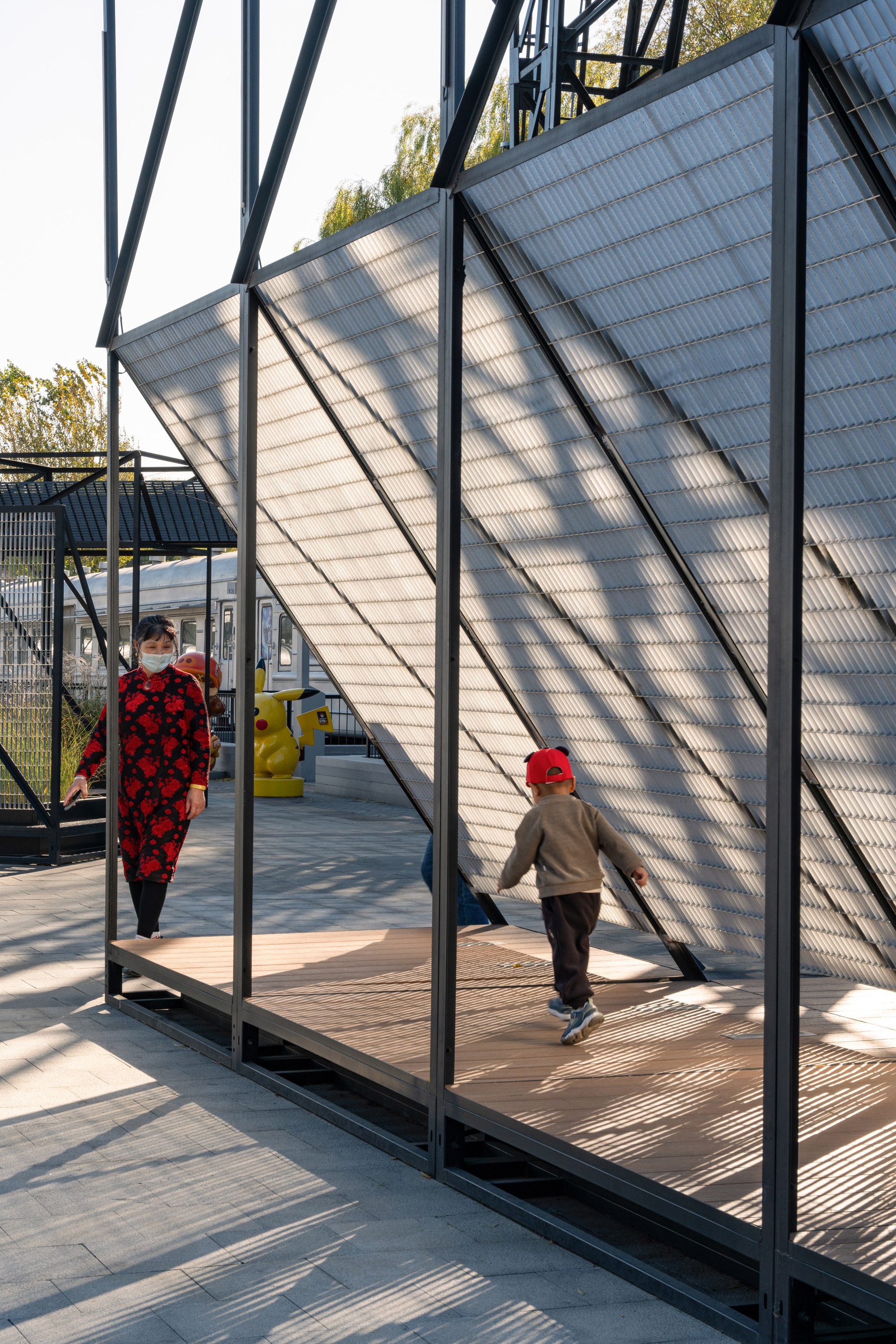 Child in a red cap runs across a timber deck beneath a slanted mesh screen casting striped shadows