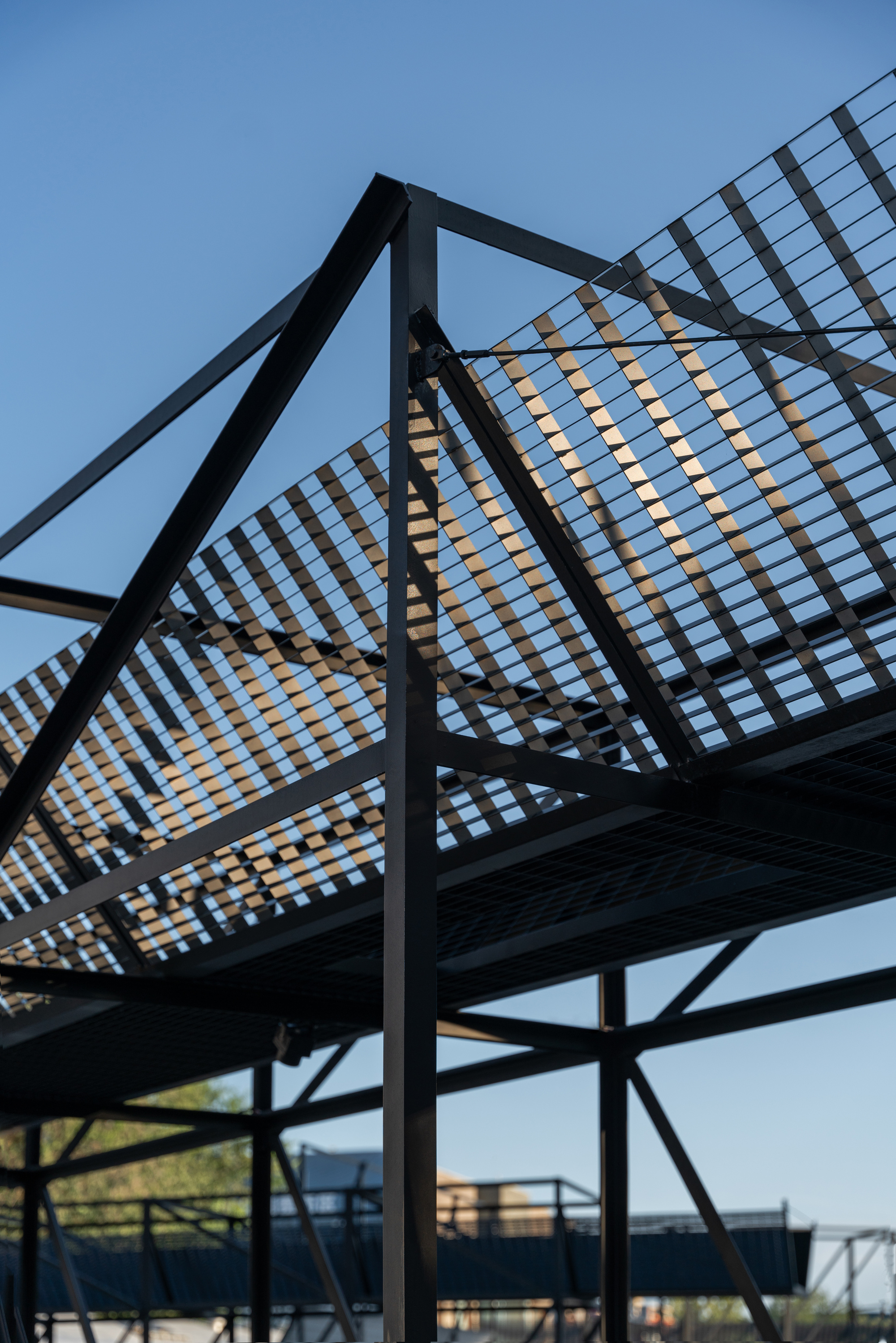 Upward view of black steel frame with slatted grating roof planes, backlit against a clear blue afternoon sky