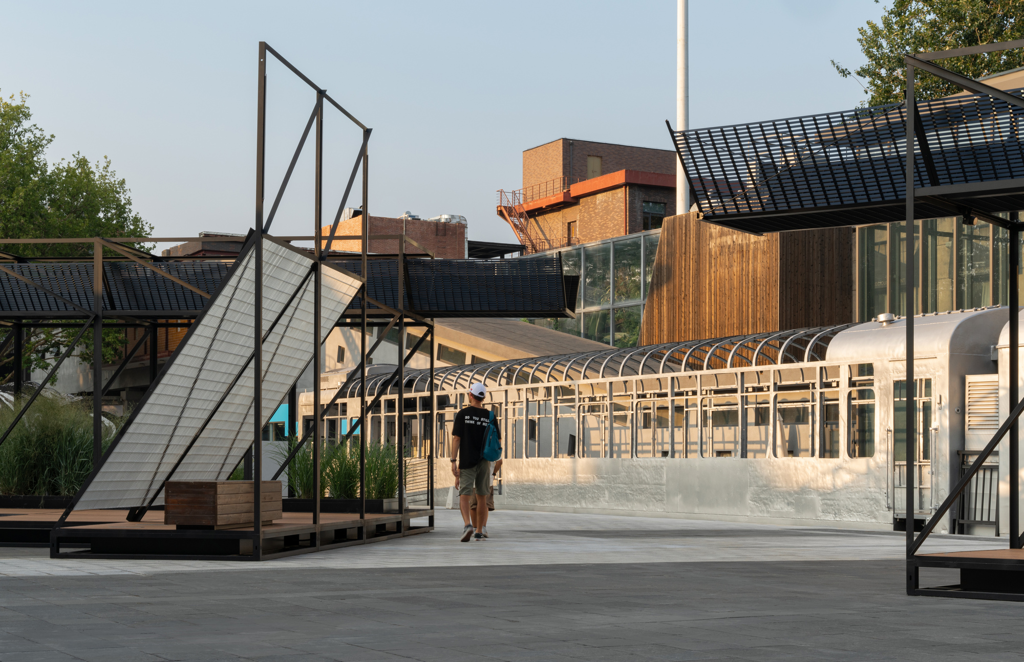 Pedestrian walking past steel frame pavilions with grating canopies and a silver railway carriage in warm late light