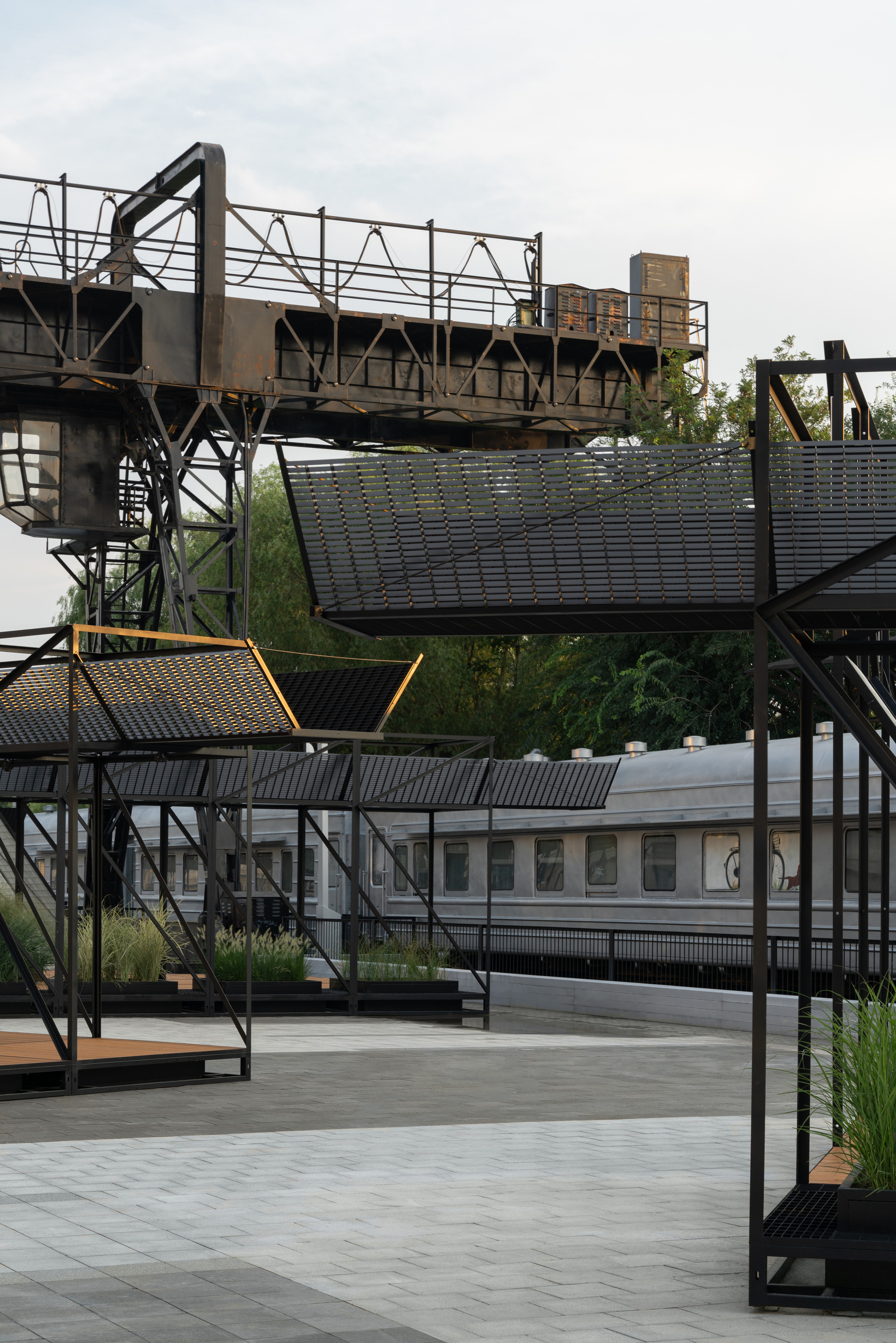 Black steel canopies and planter boxes in front of a parked train car and a rusted gantry under overcast sky