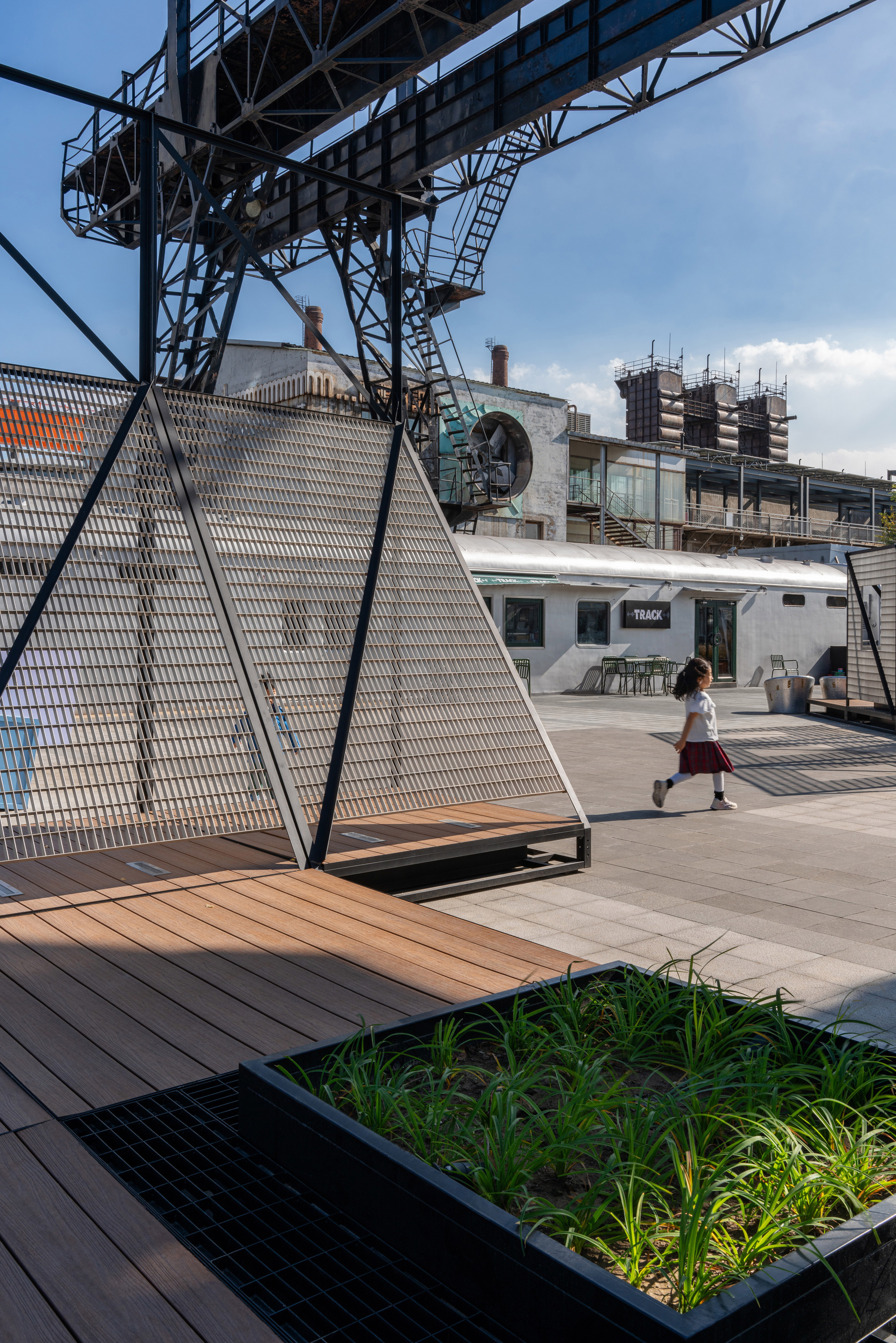 Angled mesh panel on a timber deck beside a grass planter, with a child running across the paving behind