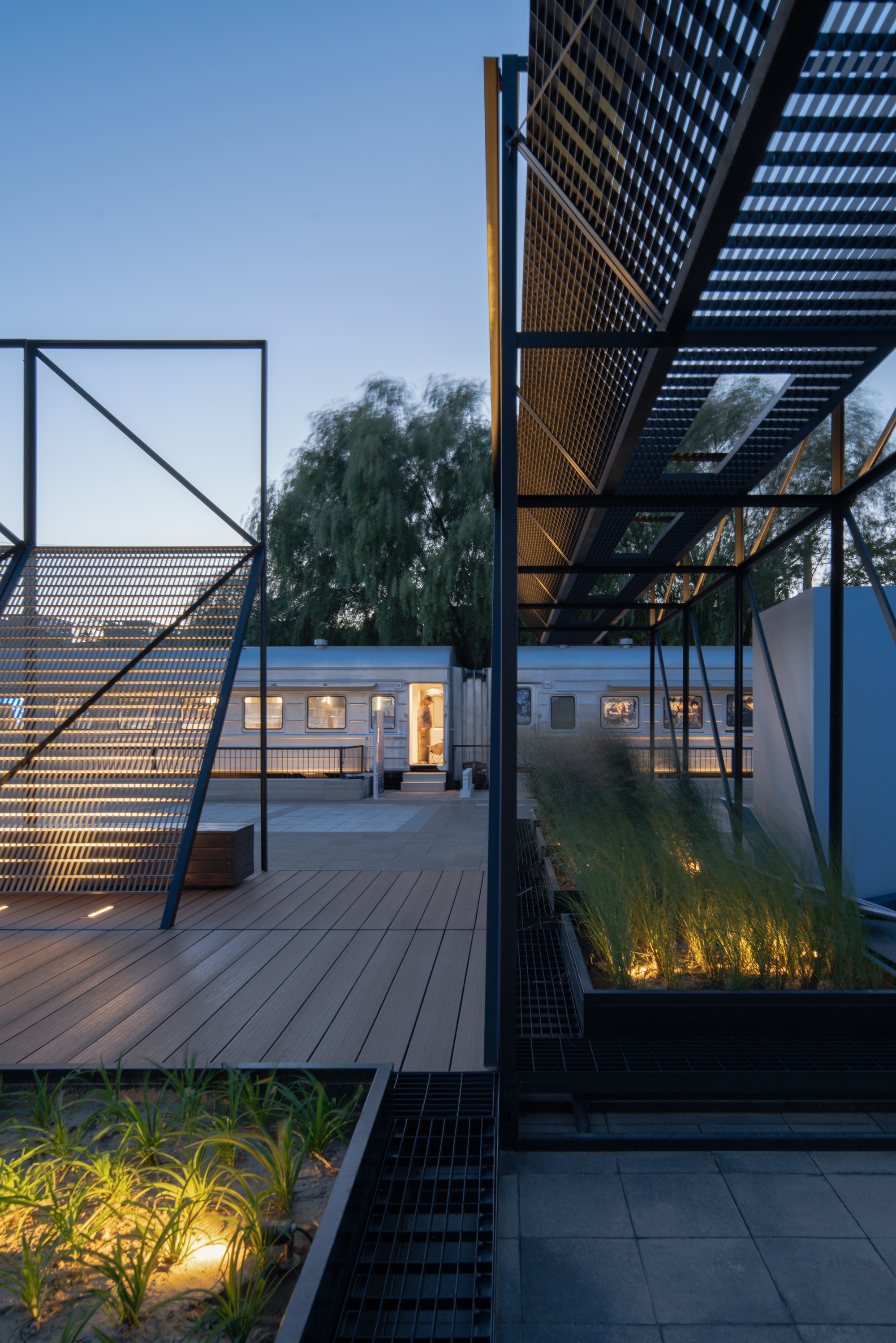 Dusk view across a timber deck with lit grass planters and a glowing train carriage doorway beyond the steel frames