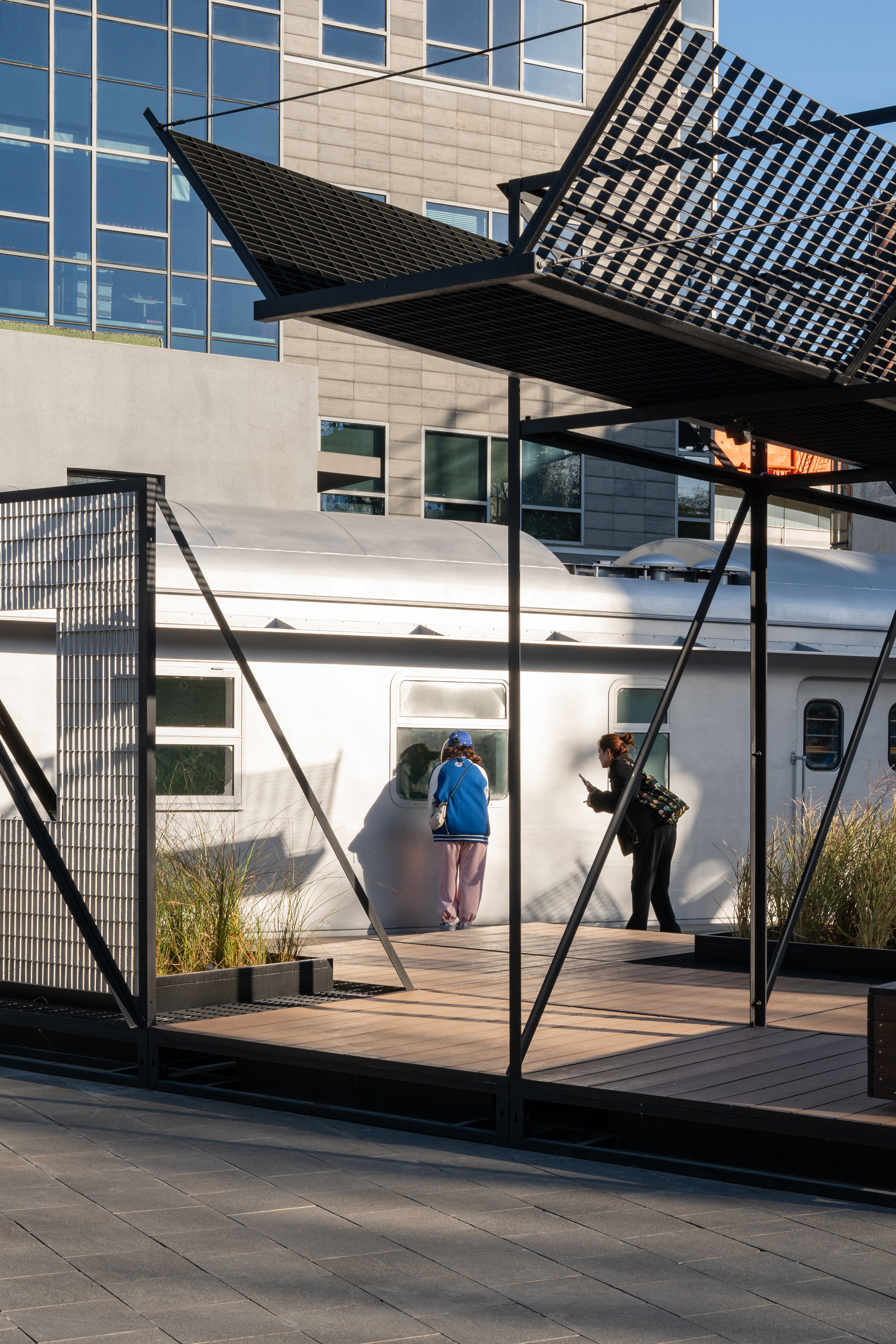 Two visitors peering into a silver train carriage window from a timber platform beneath a tilted grating canopy in low sun