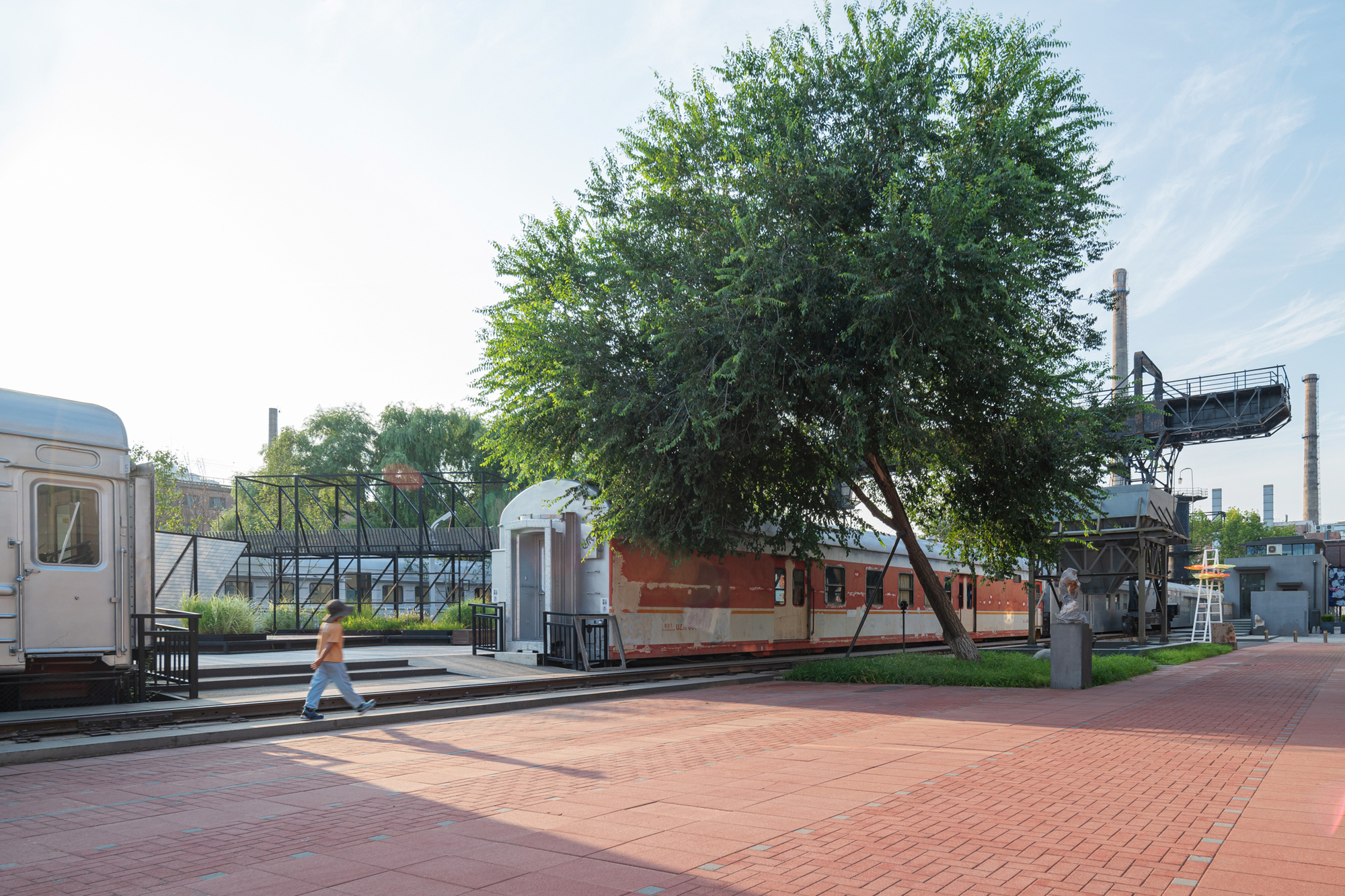 Weathered red and silver railway carriages beside a leaning tree on a brick-paved plaza, a pedestrian crossing in warm afternoon sun