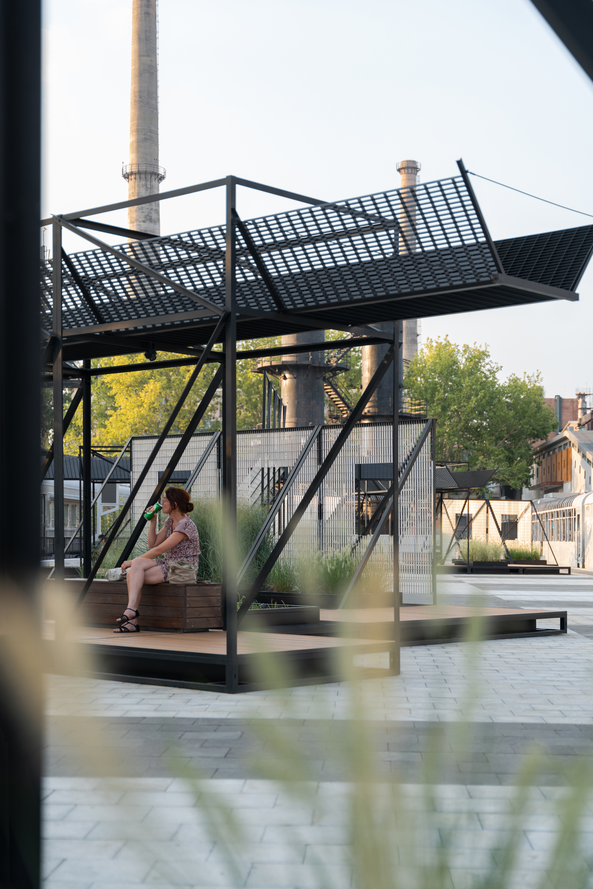 Black steel frame pavilion with a mesh canopy and timber platform, a seated woman drinking beneath industrial chimneys