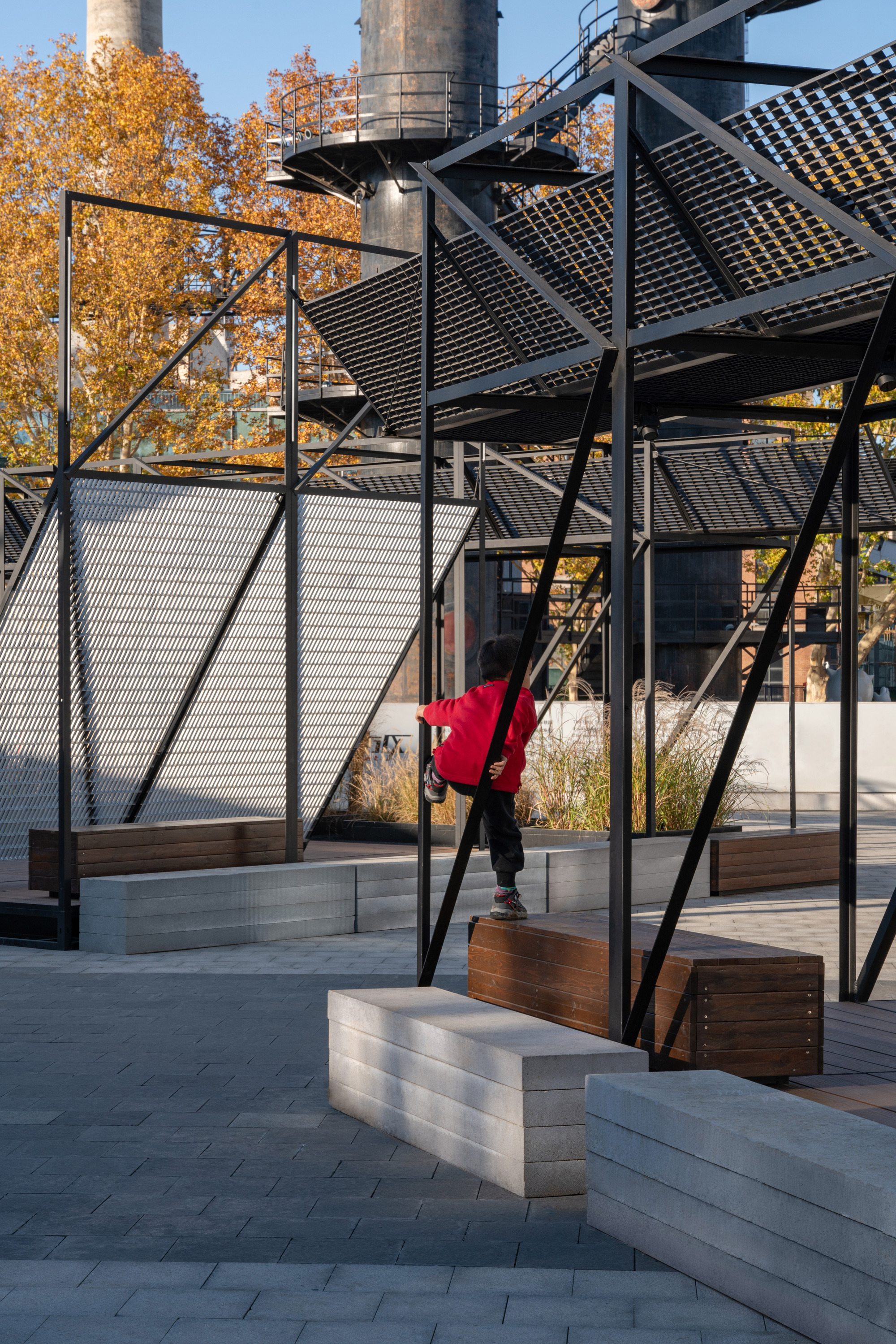 Child in a red jacket climbing a diagonal steel brace above concrete and timber benches, with yellow autumn trees behind