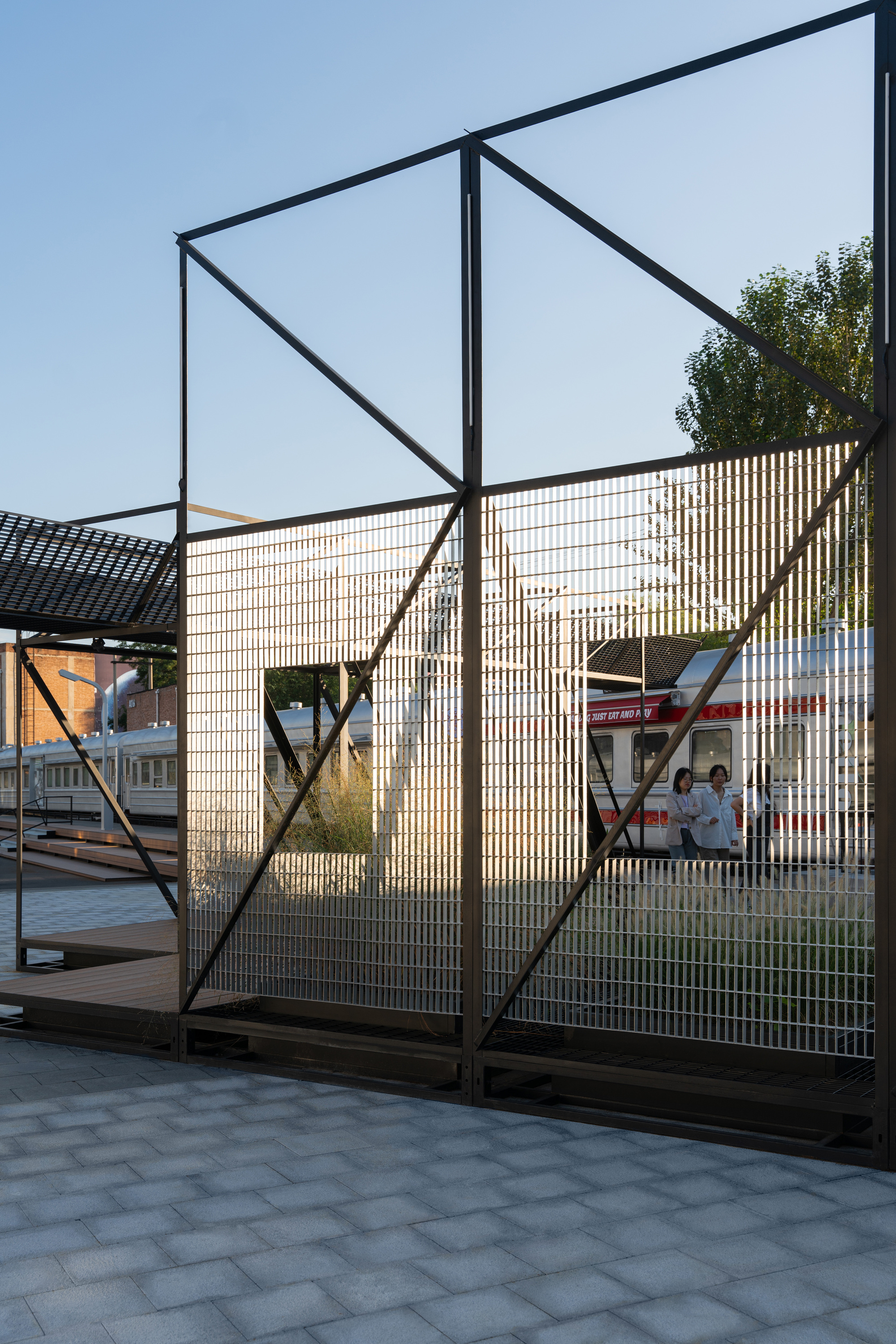 Steel frame with diagonal bracing and grating panels, framing a view of railway carriages and visitors at low evening sun