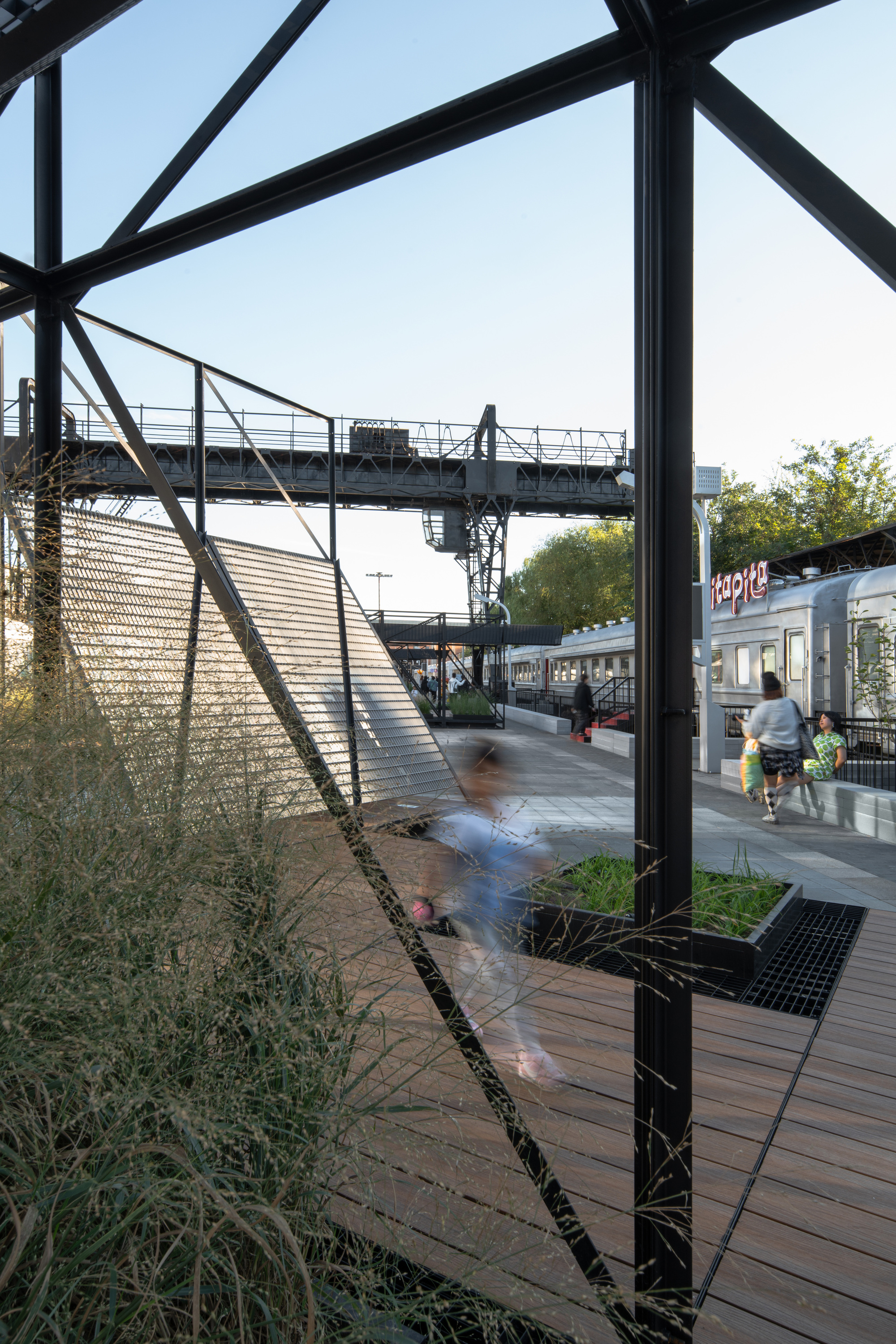 Blurred figure running across a timber deck past planter beds and grasses, with a steel footbridge and train carriage beyond
