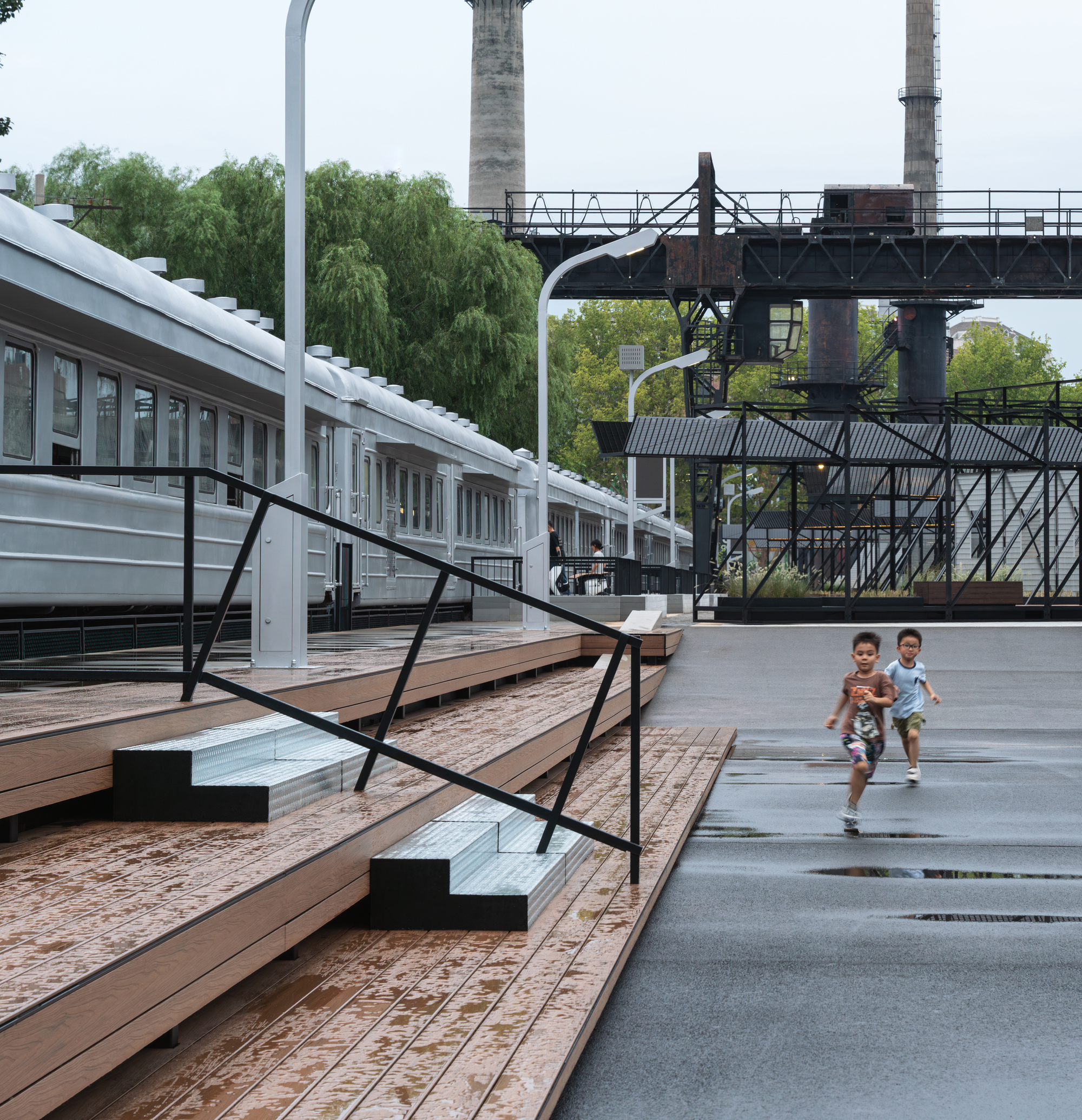Timber deck steps with metal risers beside a silver railway carriage, two children running across wet paving under an overcast sky
