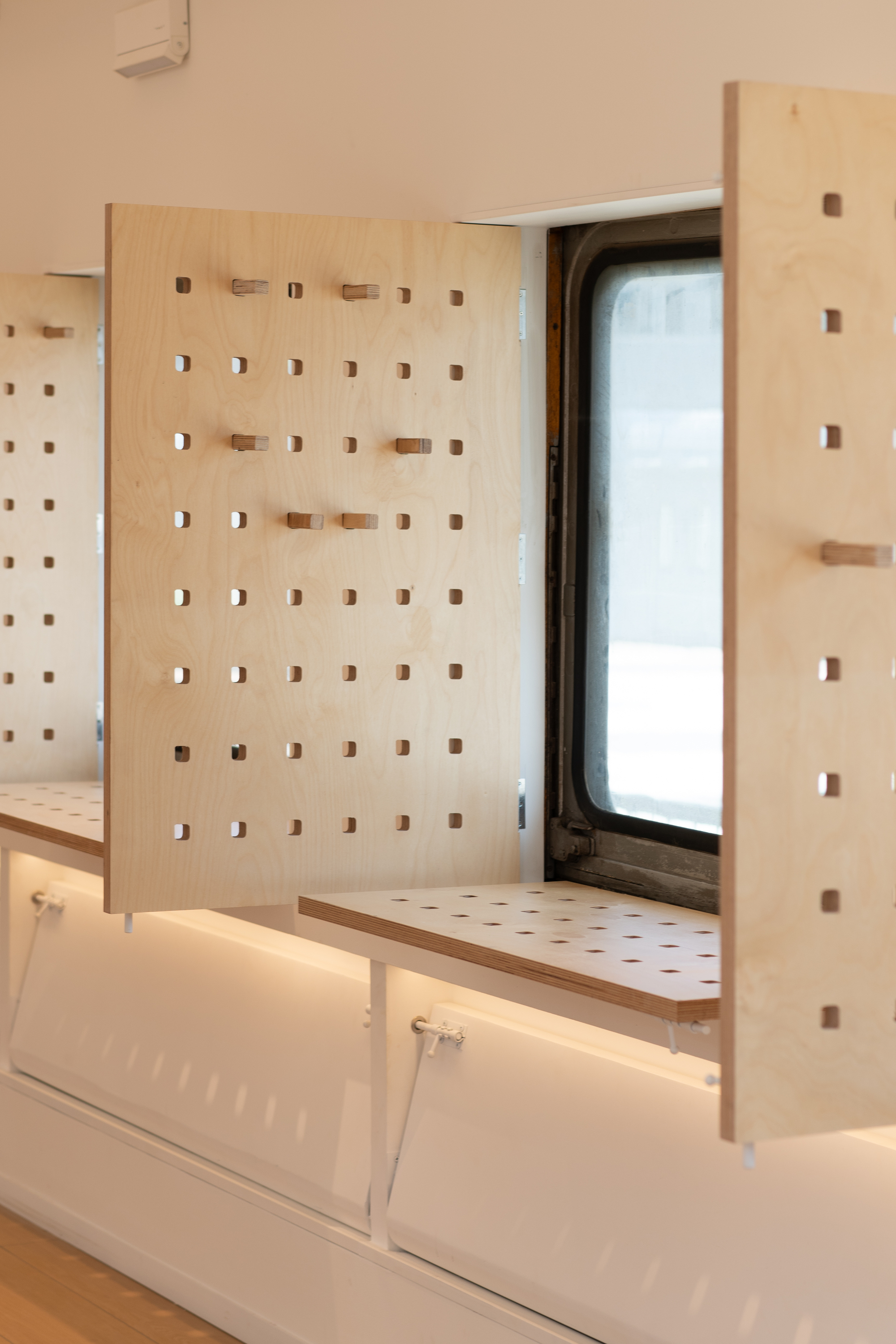 Close-up of perforated birch plywood shutters opened beside a carriage window, with a fold-down shelf and lit storage below