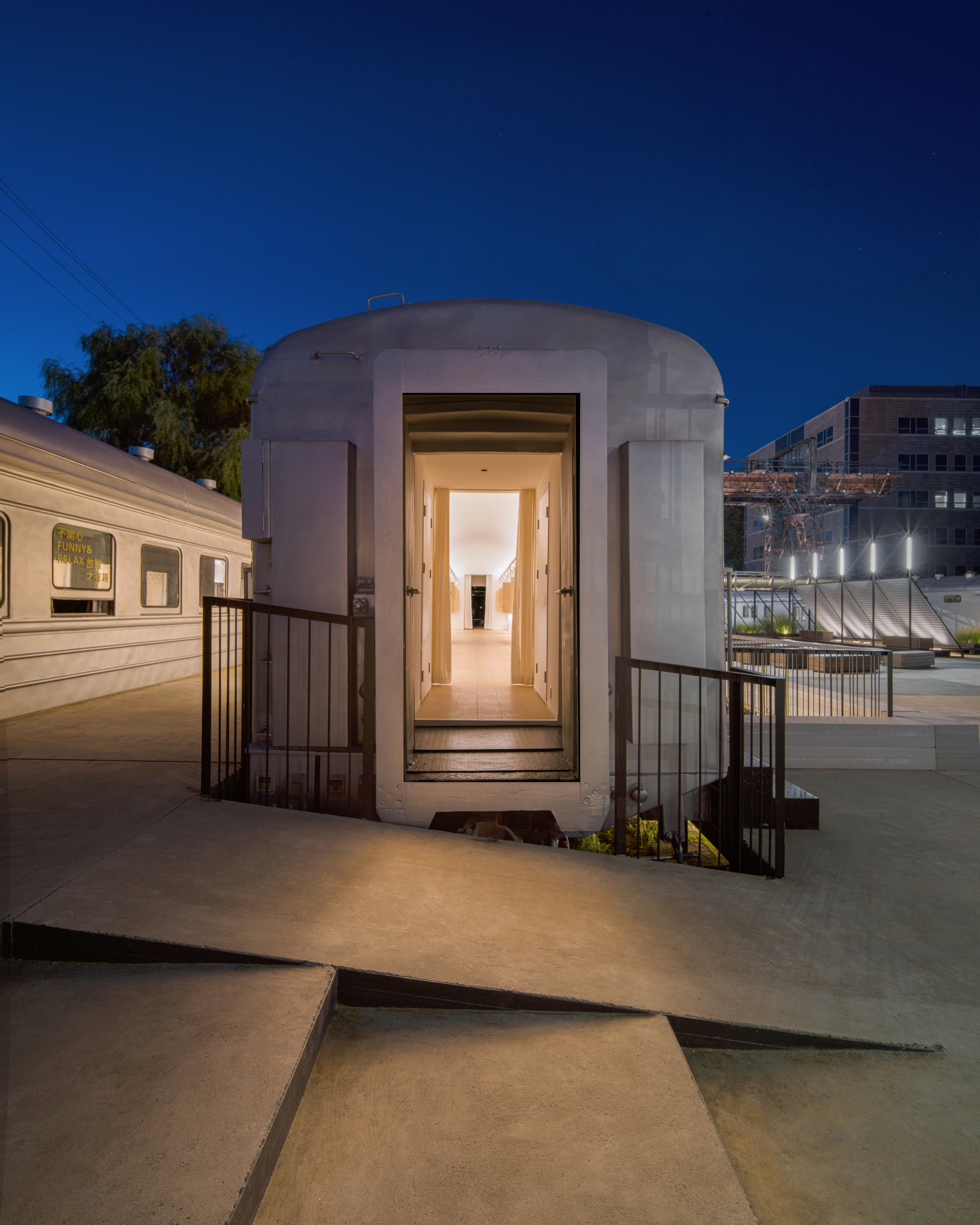 Rail carriage end with an open glowing doorway, railings and angled concrete ramps under a deep blue dusk sky