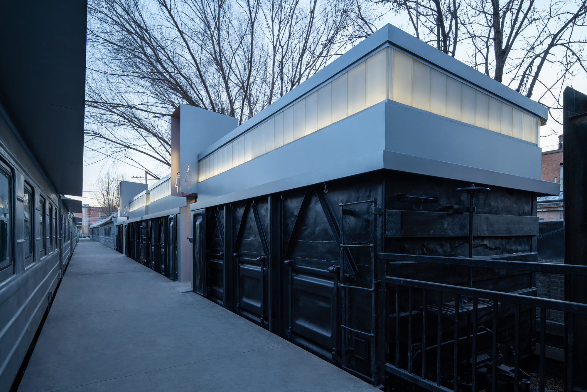Narrow walkway between a train and black freight wagons topped by a translucent glowing band, with bare trees overhead