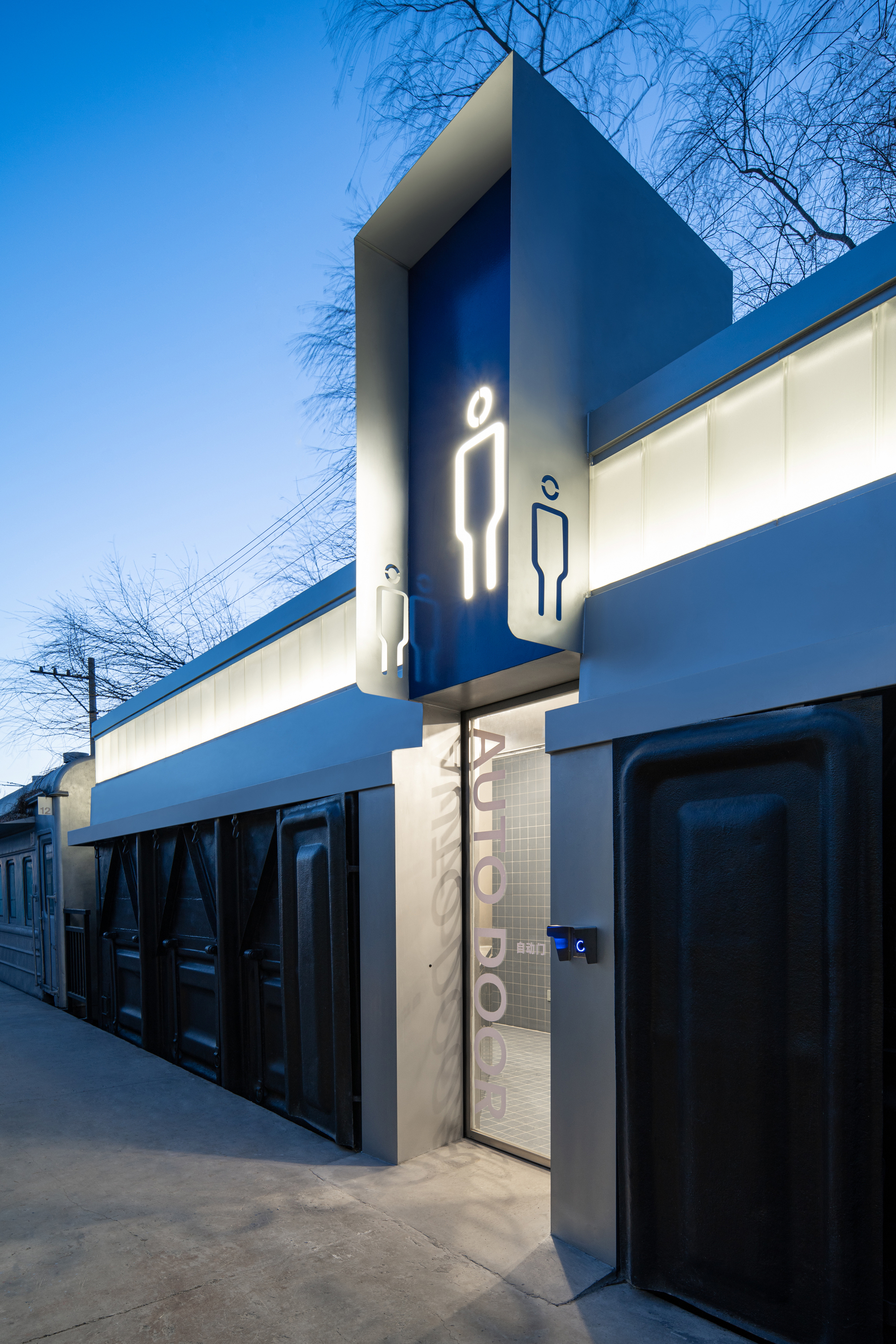Entrance to a public restroom with a glowing illuminated male pictogram on a blue panel, dark ribbed doors and an automatic door at dusk