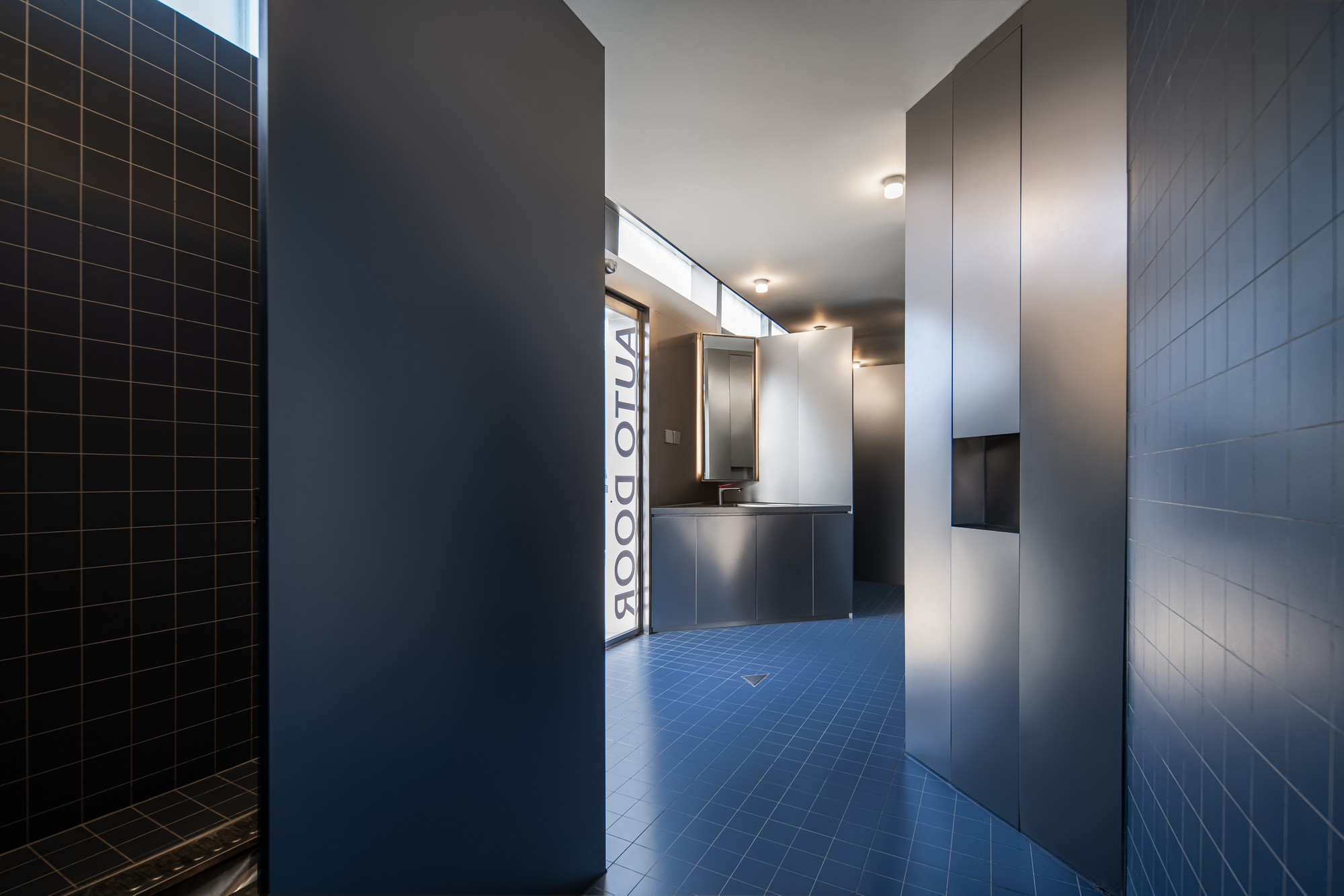 Blue-tiled corridor leading toward a steel vanity and a glass door printed with AUTO DOOR, with clerestory light above