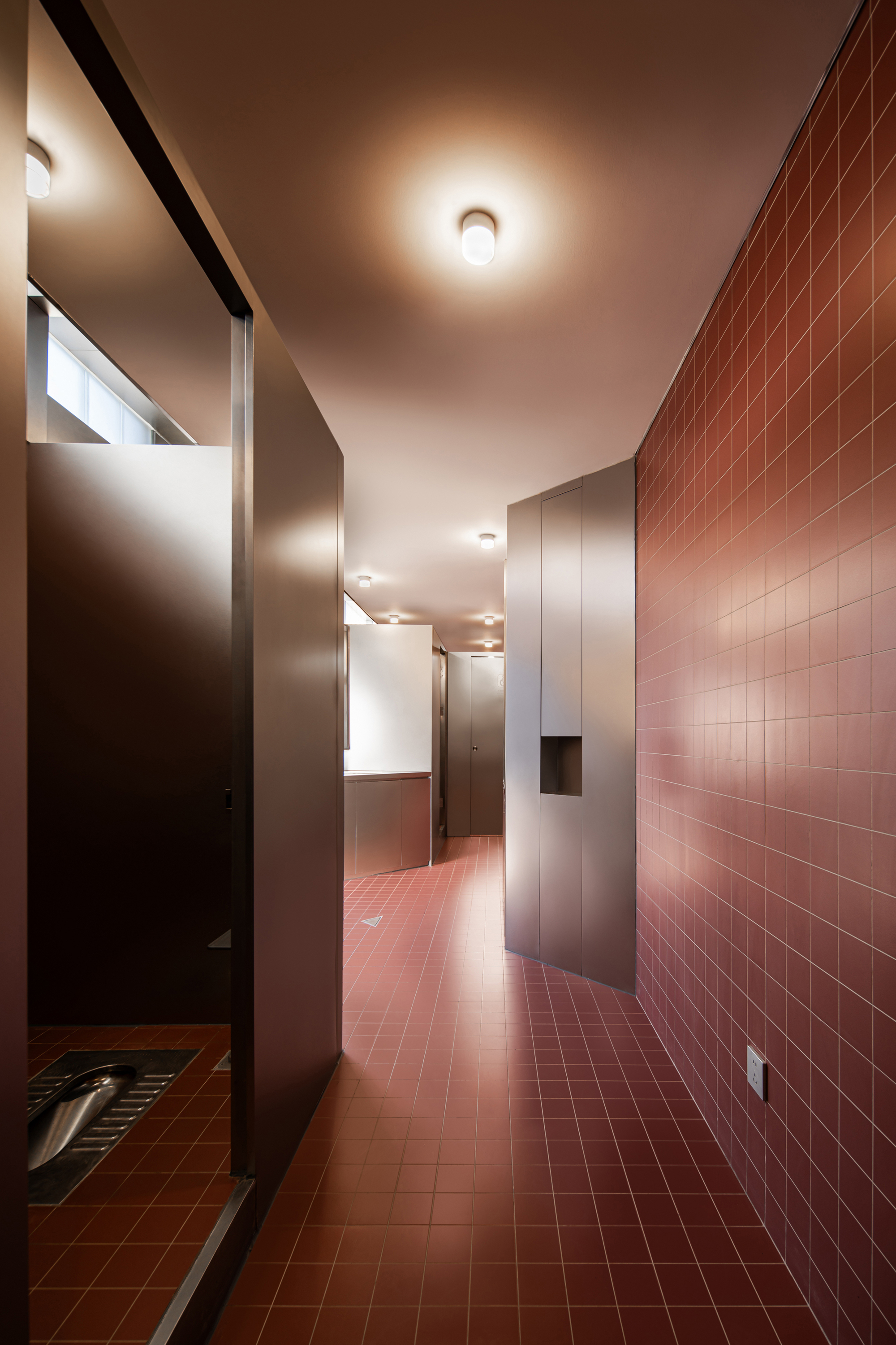 Red-tiled restroom corridor with a squat toilet stall at left, a steel storage niche and ceiling lamps glowing along the passage