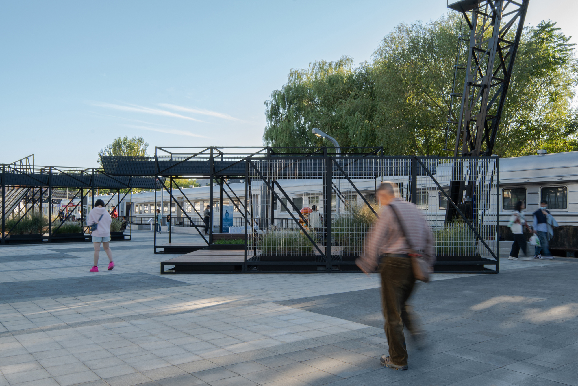 Black steel frame pavilions with mesh panels and planted beds beside a parked train carriage, with a blurred man walking past in daylight