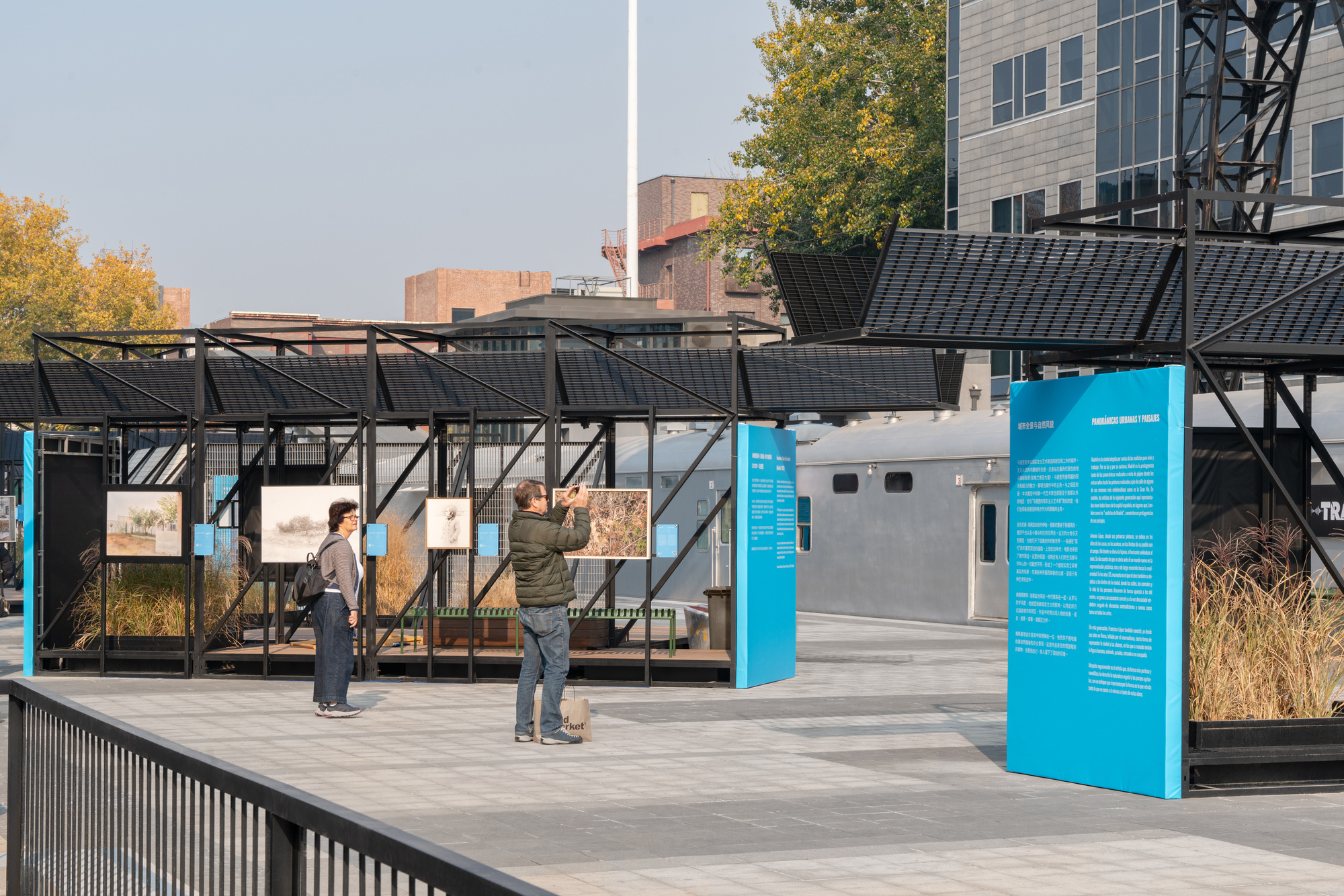 Visitors viewing framed photographs inside black steel exhibition pavilions, with tall cyan text panels and autumn trees in the background