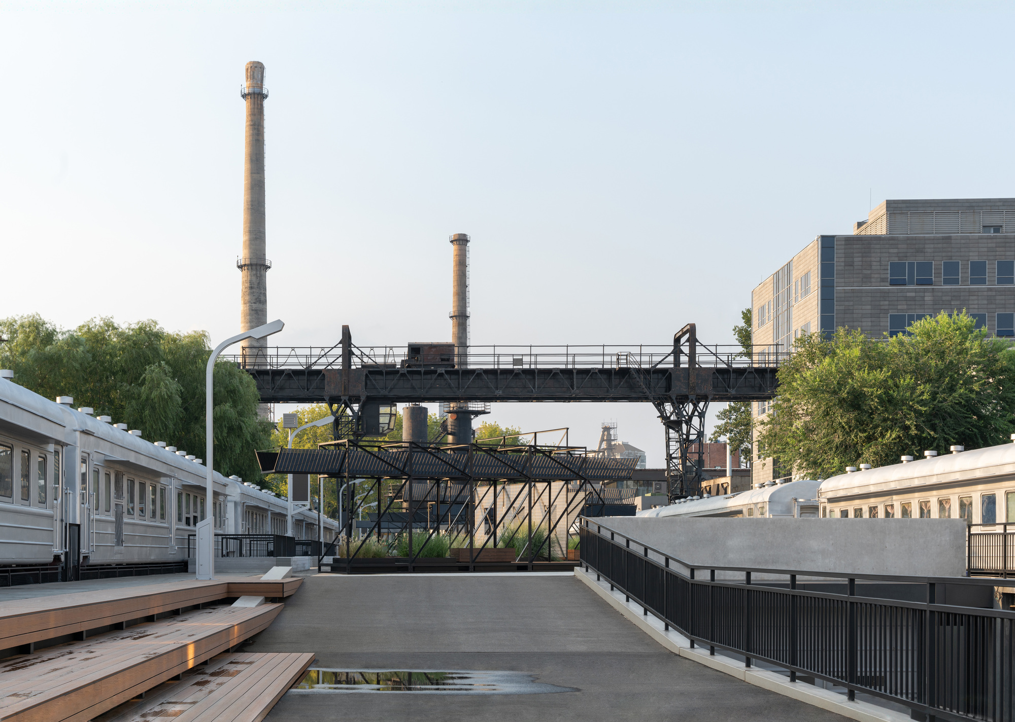 Black steel gantry bridge and smokestacks beyond a concrete ramp, timber steps and railway carriages in clear morning light