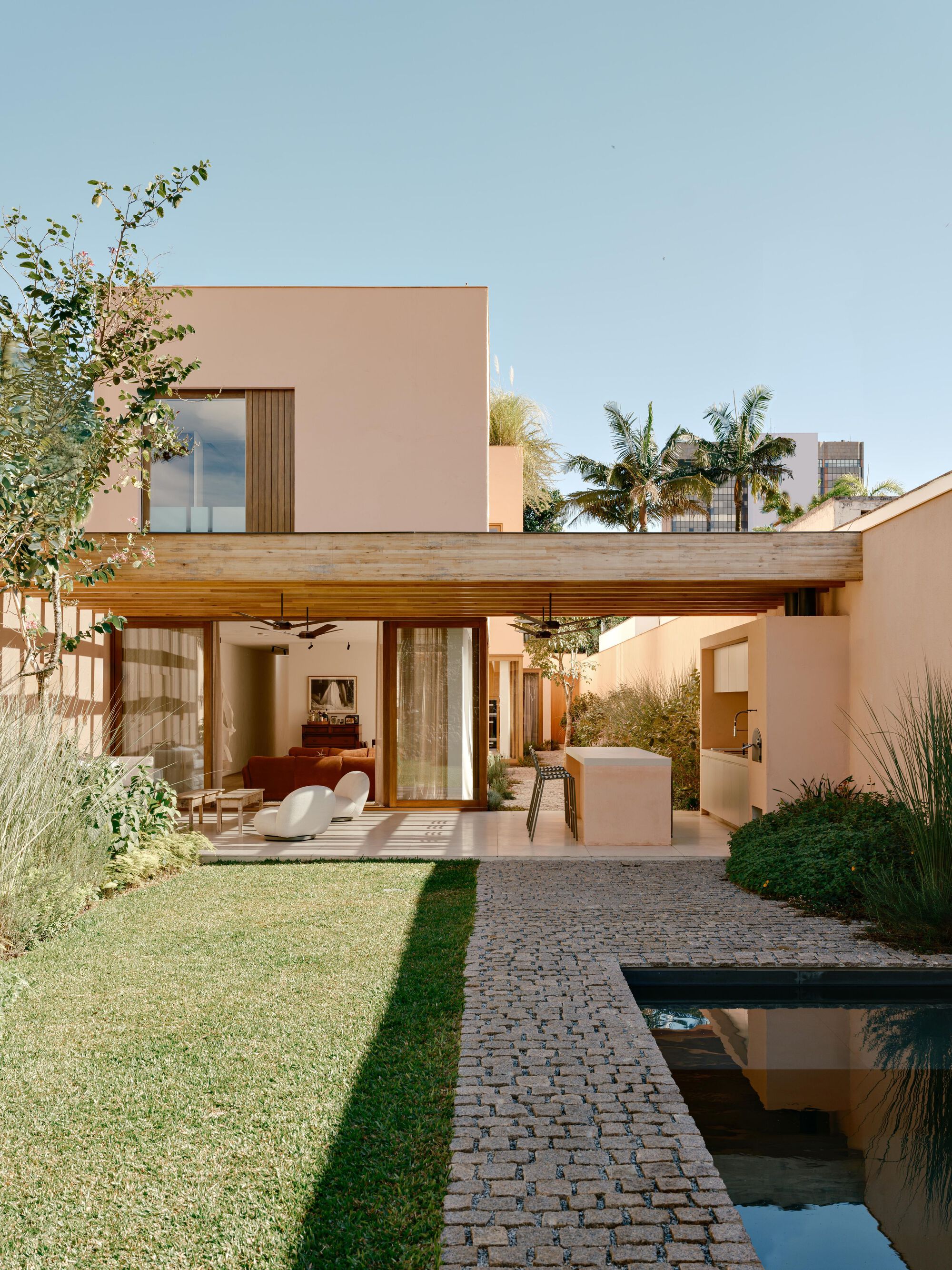 Garden facade with a timber-soffit canopy over an open living room, a cobbled path and reflecting pool in clear midday sun