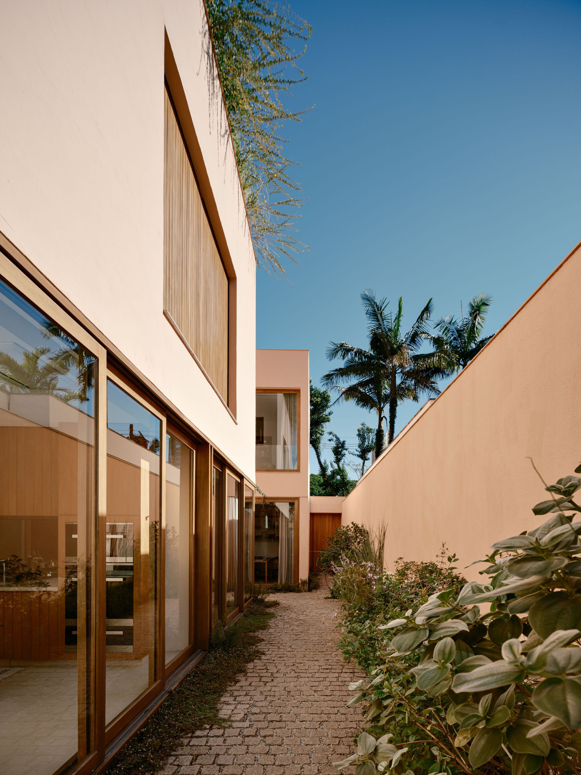 Narrow side passage of cobblestones between a glazed timber-framed wall and a pink boundary wall, with palms against a blue sky