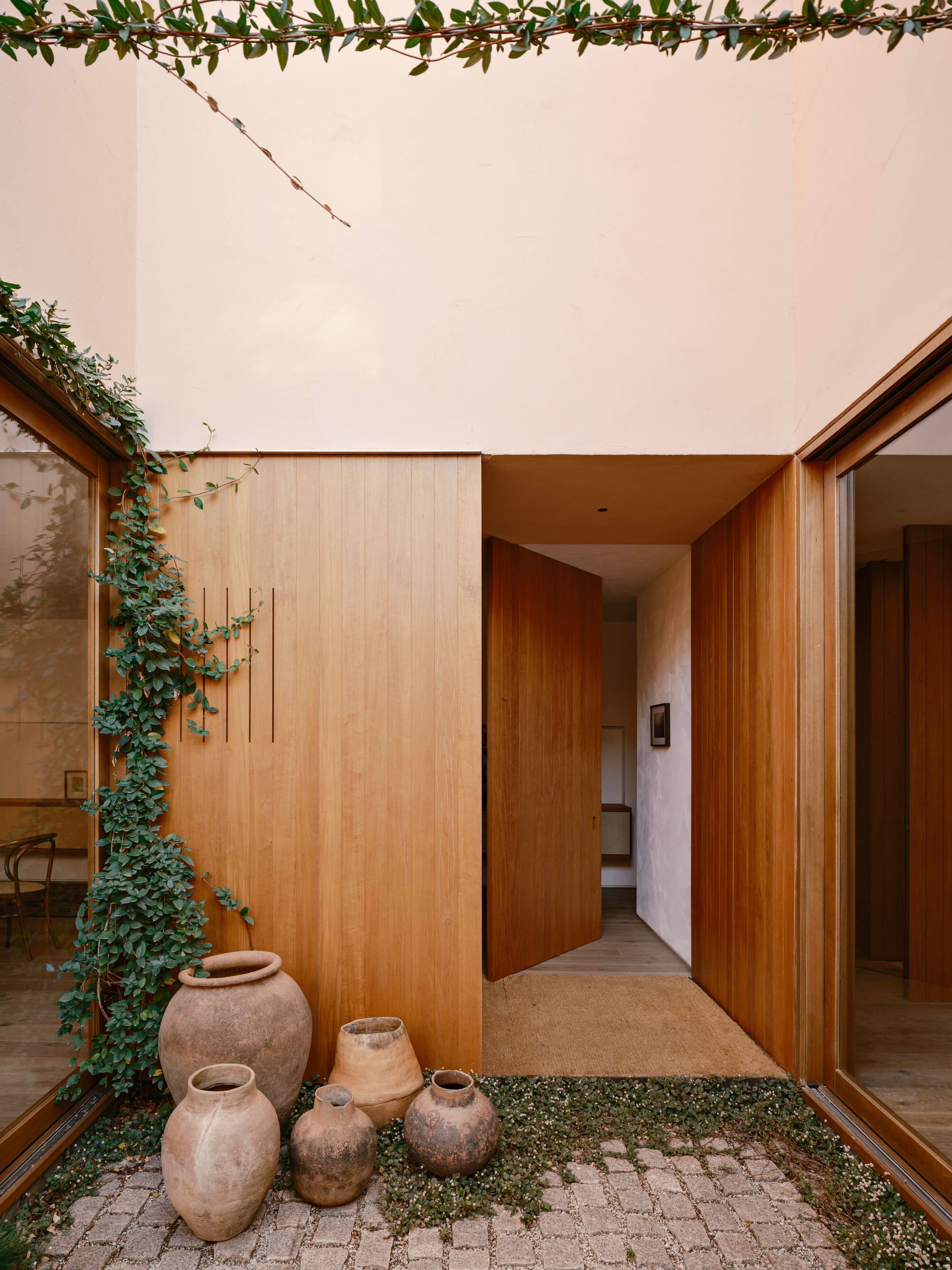 Courtyard entry with a pivot timber door, clay jars on a pebble and moss bed, and a climbing vine on the wall