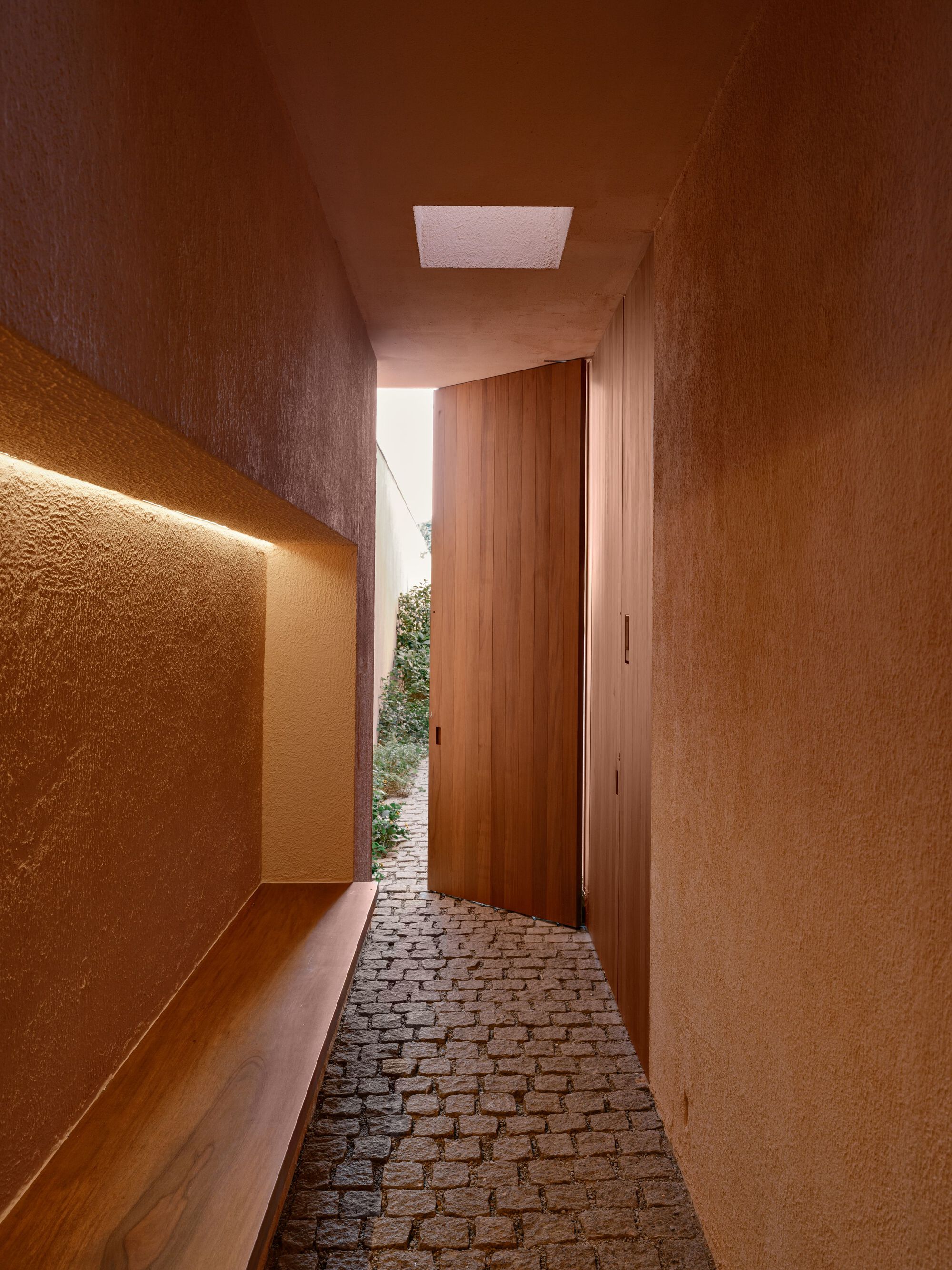 Textured plaster corridor with a lit built-in timber bench, a skylight and an open pivot door onto a cobbled path