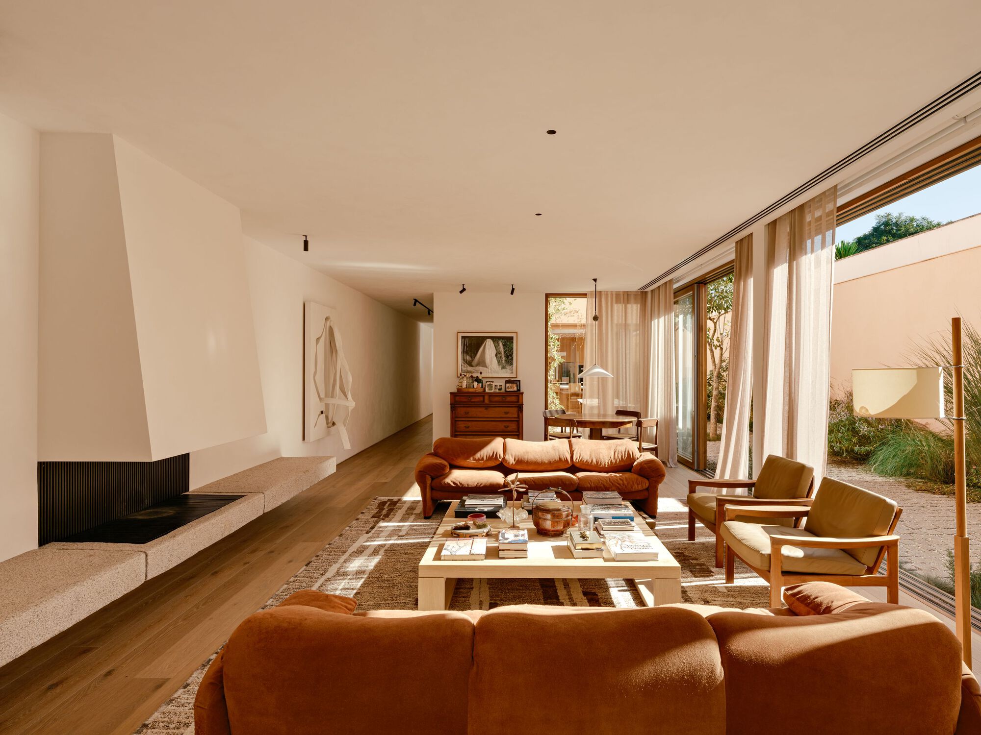 Long living space with a plaster fireplace and stone ledge, sunlit rug and armchairs, and sliding glass doors opening to the courtyard
