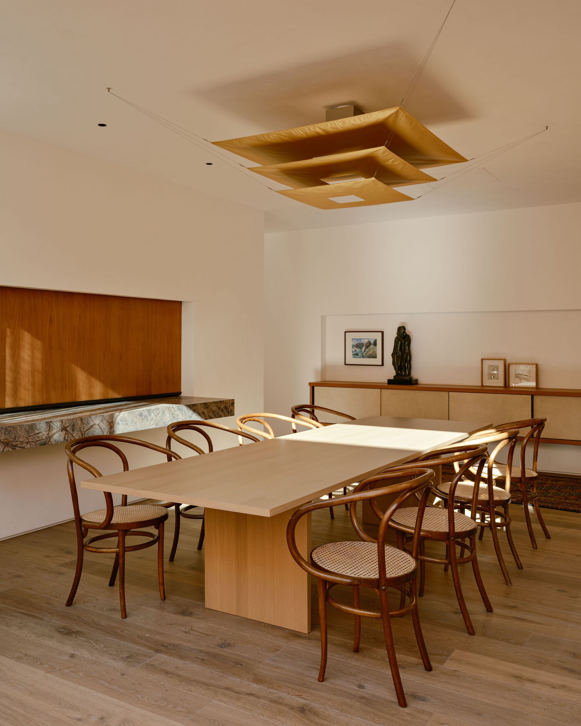 Dining room with oak table, bentwood cane chairs and a layered golden ceiling pendant, with sunlight crossing the tabletop