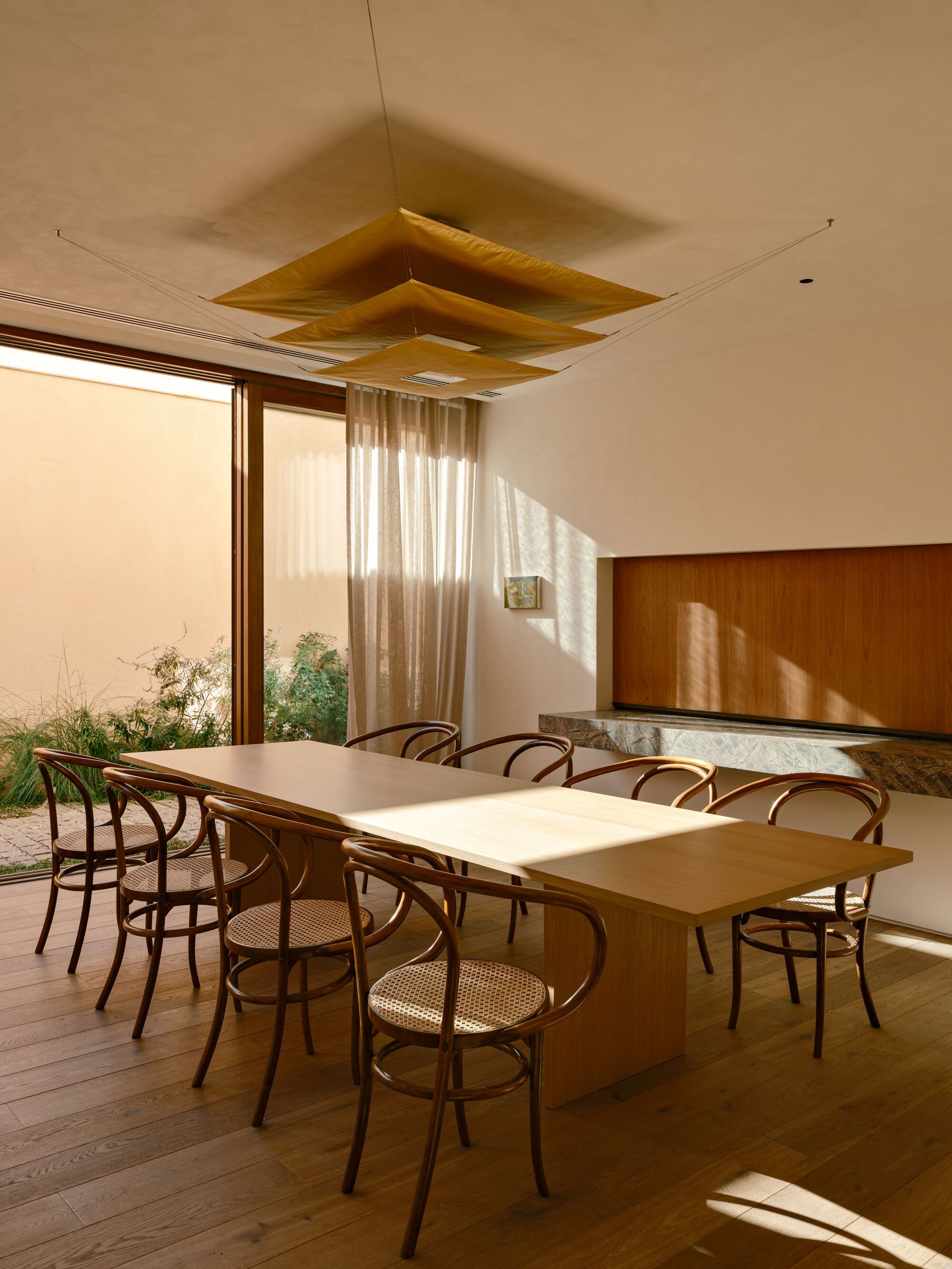 Long oak dining table and bentwood chairs beside a glazed wall opening to a planted courtyard, with late sun on the floor