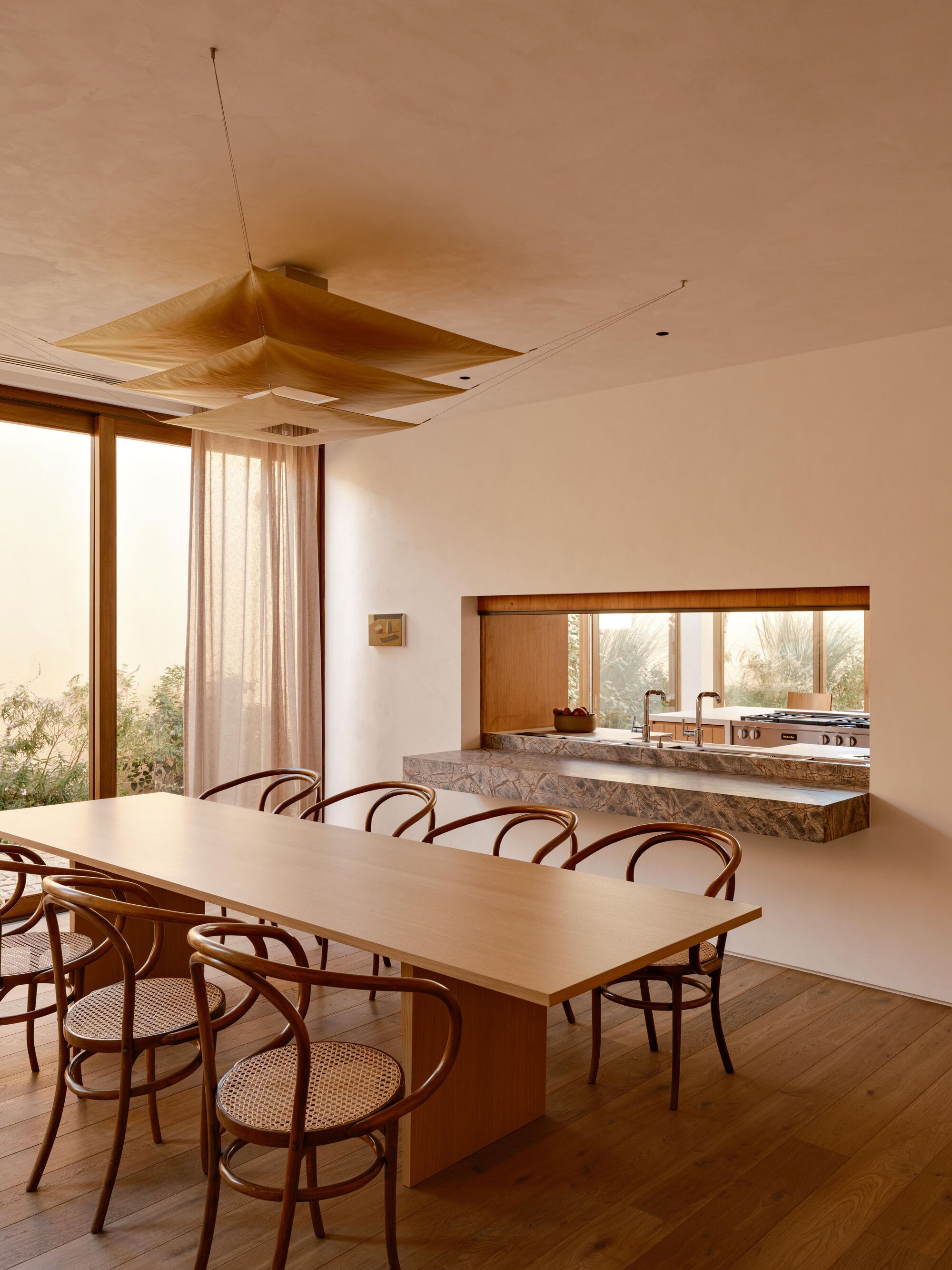 Dining table facing a stone-edged pass-through to the kitchen, with sheer curtains and garden glimpsed through the glass at dusk