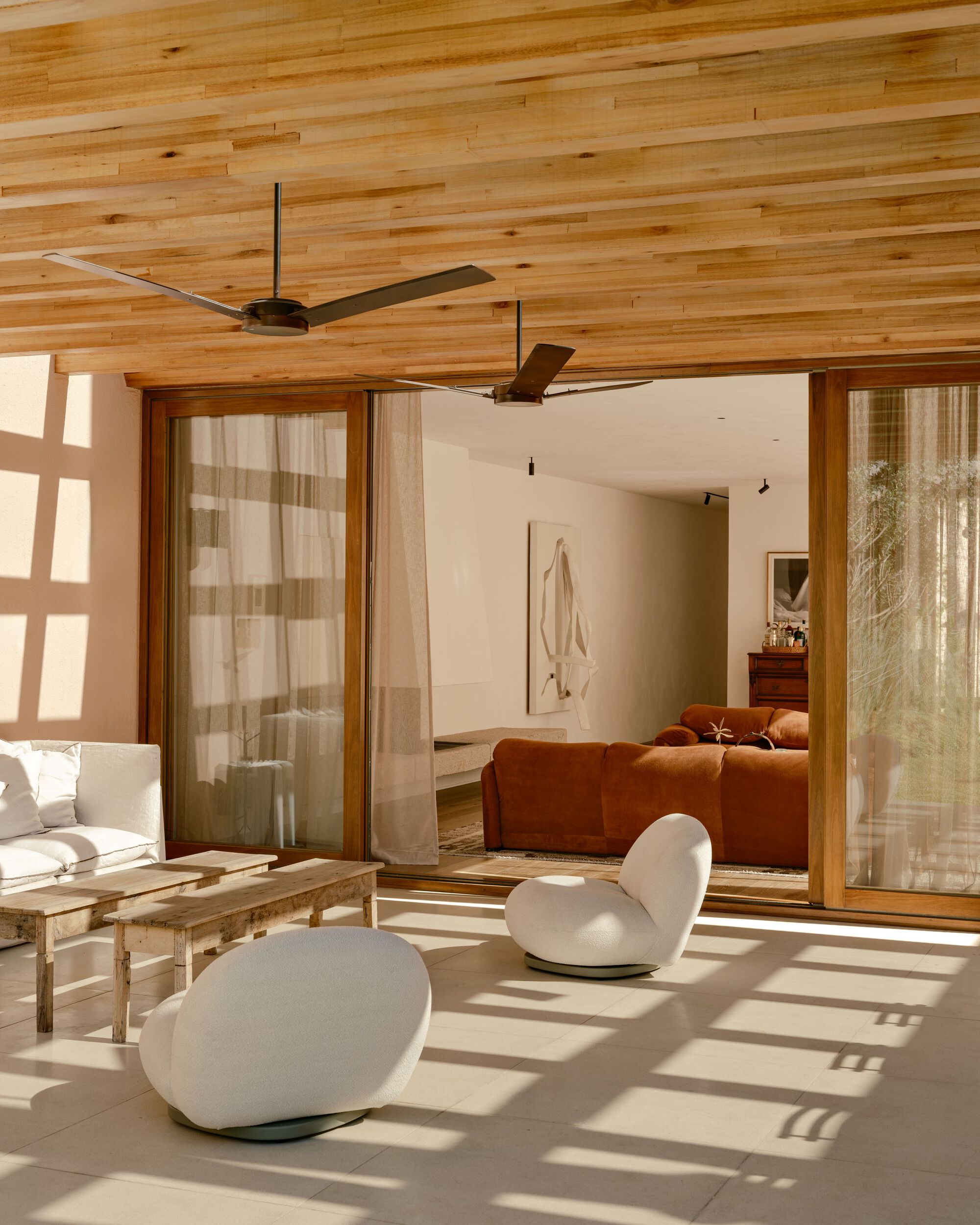 Covered terrace with timber plank ceiling, two ceiling fans and white rounded chairs, with sun patterns on the stone floor