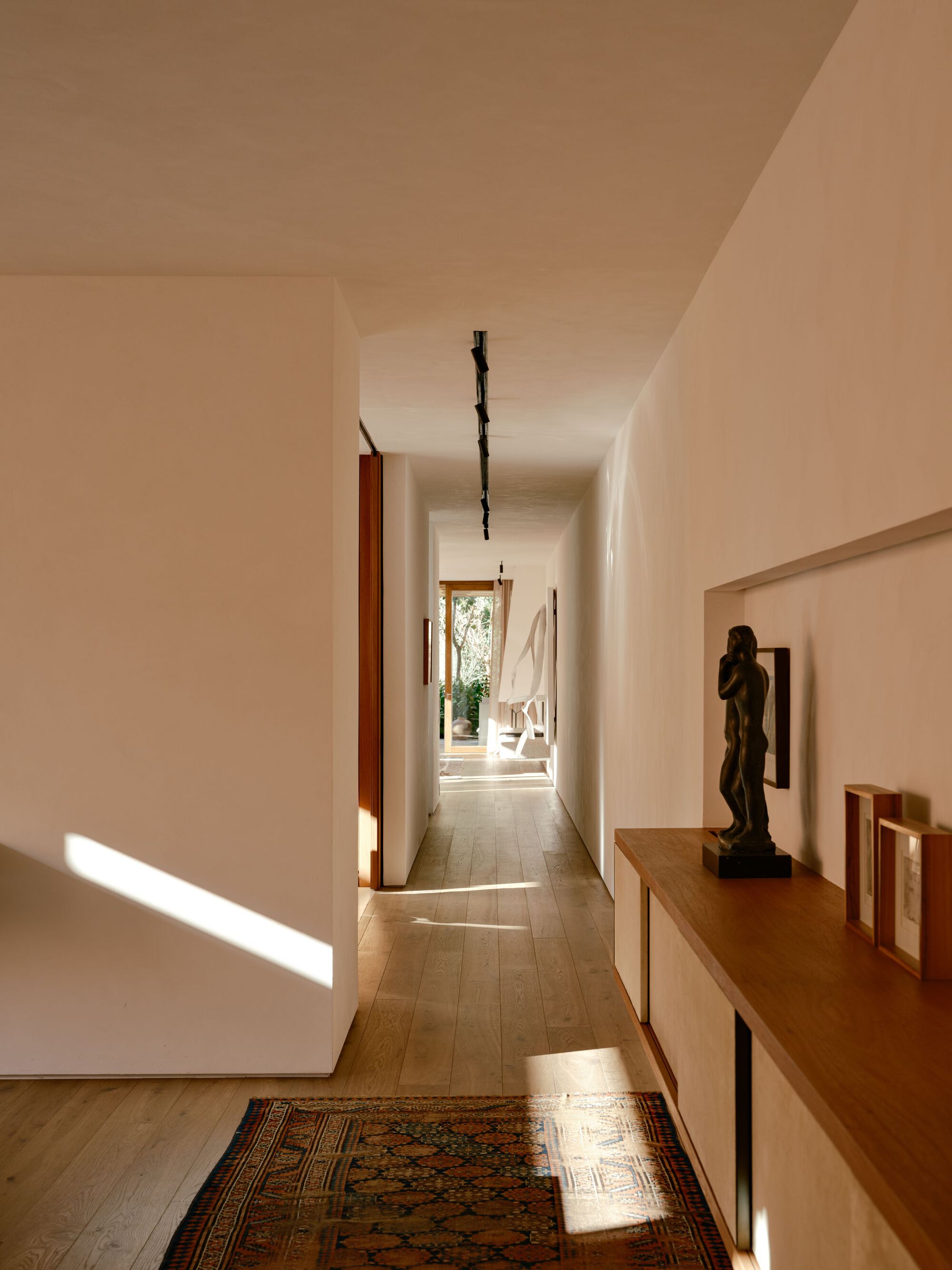 Hallway lined with a timber sideboard, bronze figure and patterned rug, with a shaft of afternoon sun on the wall