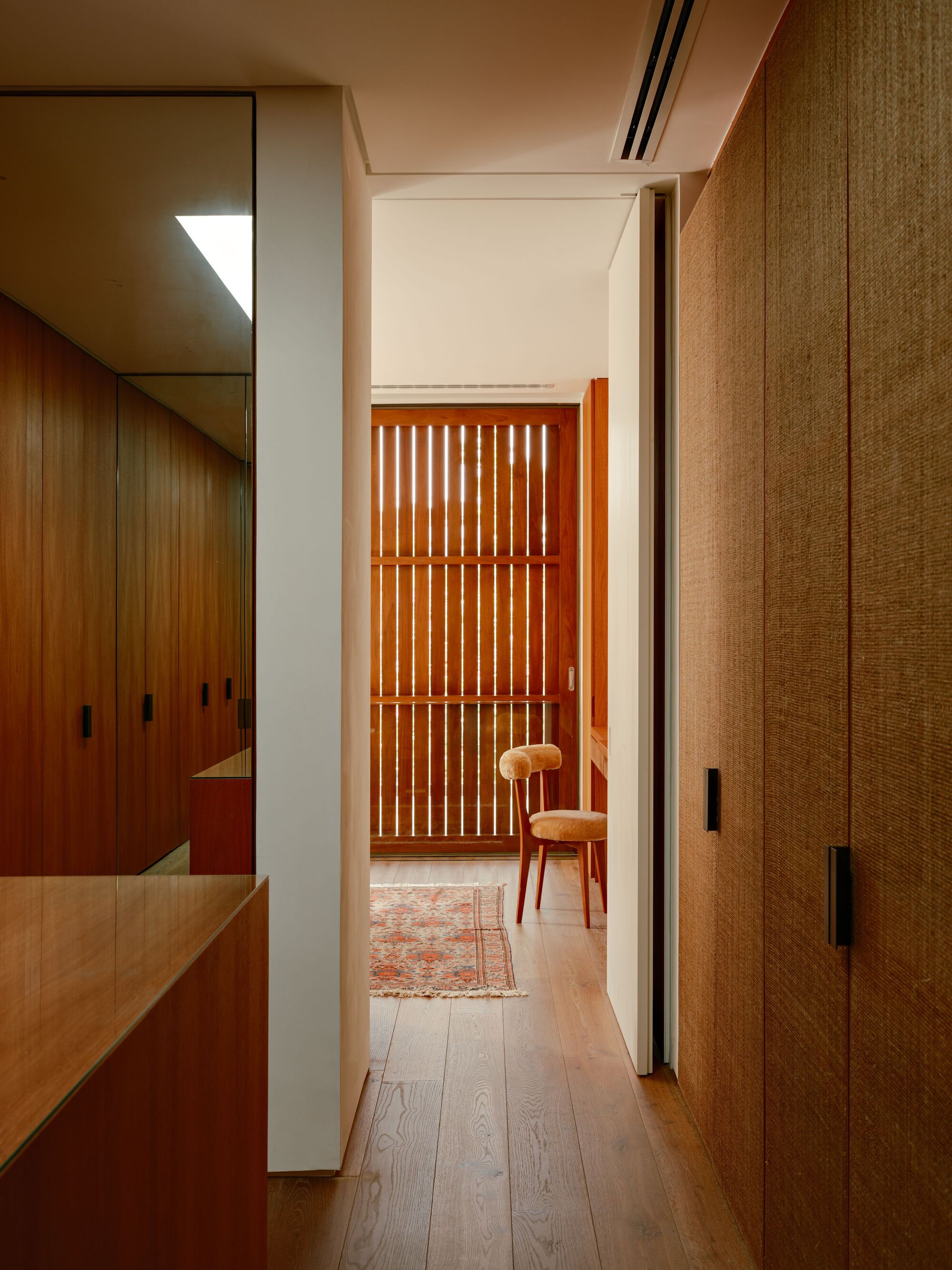 Corridor between timber joinery and woven-fibre wardrobe doors, ending at a slatted wood screen and a chair on a rug