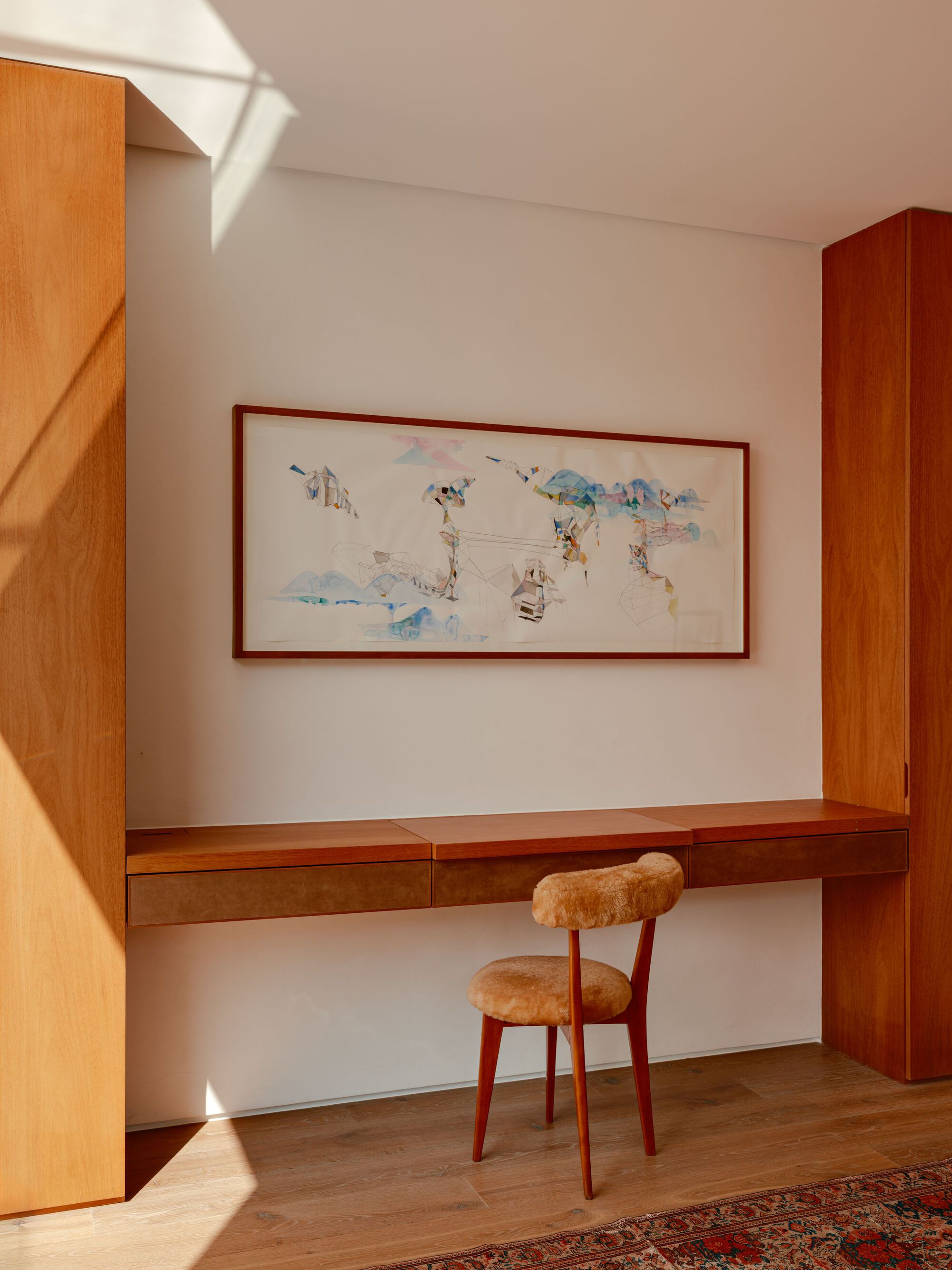 Built-in timber desk with a fur-seated chair beneath a framed watercolour, lit by a patch of low sun