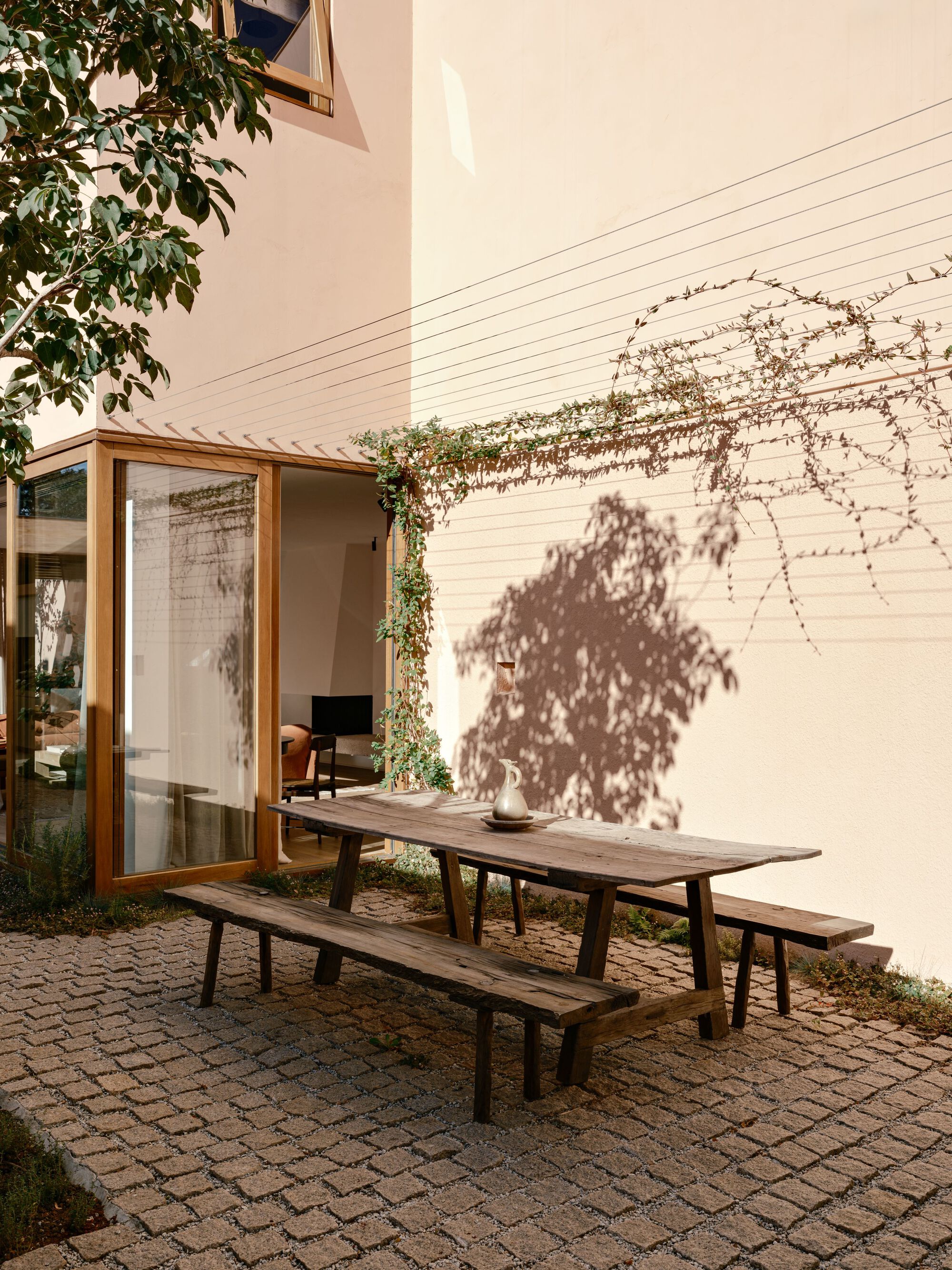 Rough timber table and benches on a cobbled patio beside a glazed corner, with vine shadows cast on the pink wall in afternoon sun