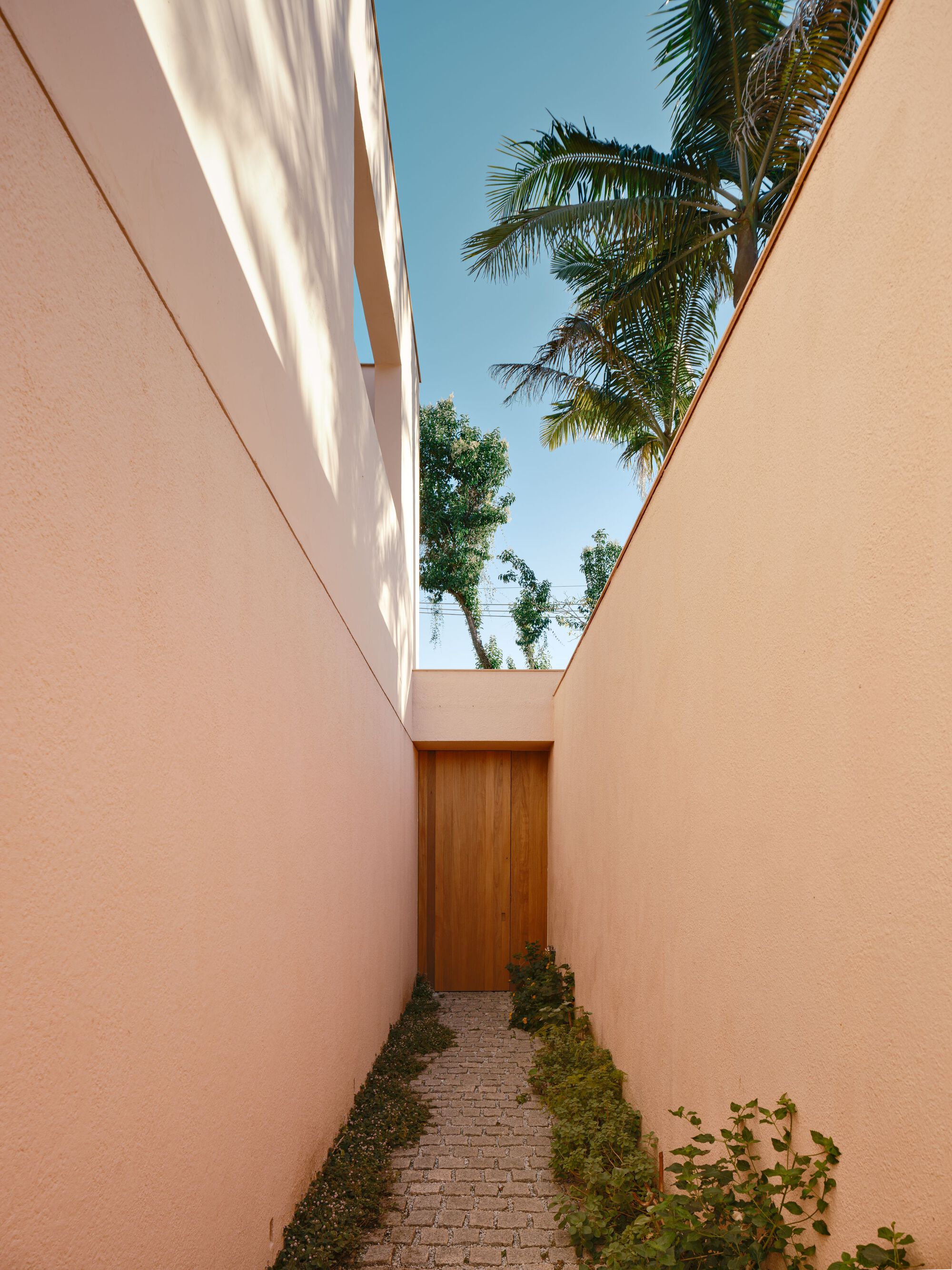 Narrow cobbled passage between tall pink walls leading to a timber gate, with palm fronds against a blue sky