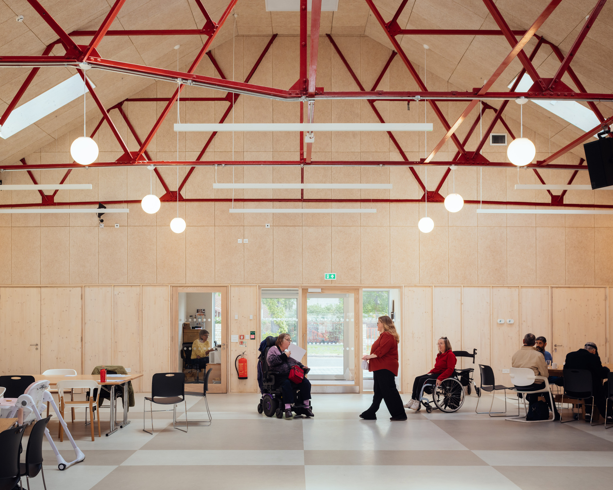 Hall interior with red steel trusses over plywood-lined walls, where people in wheelchairs and others gather at tables in daylight