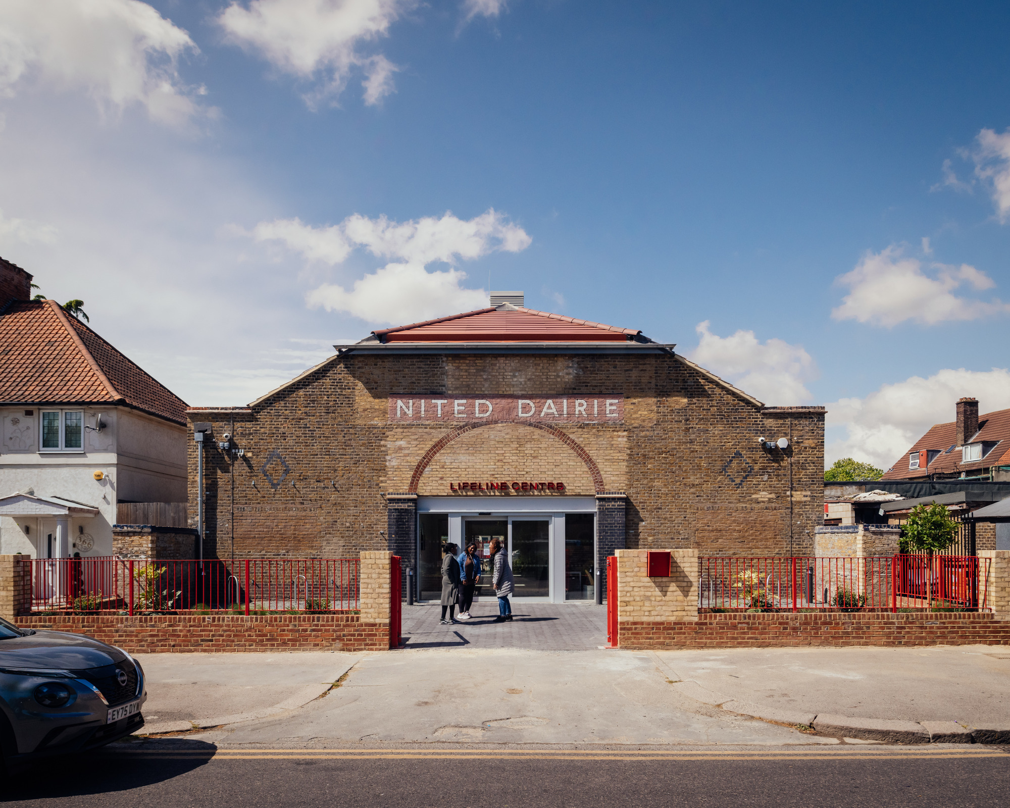 Brick street facade with arched entrance and painted lettering, red railings in front and three people talking under a blue sky