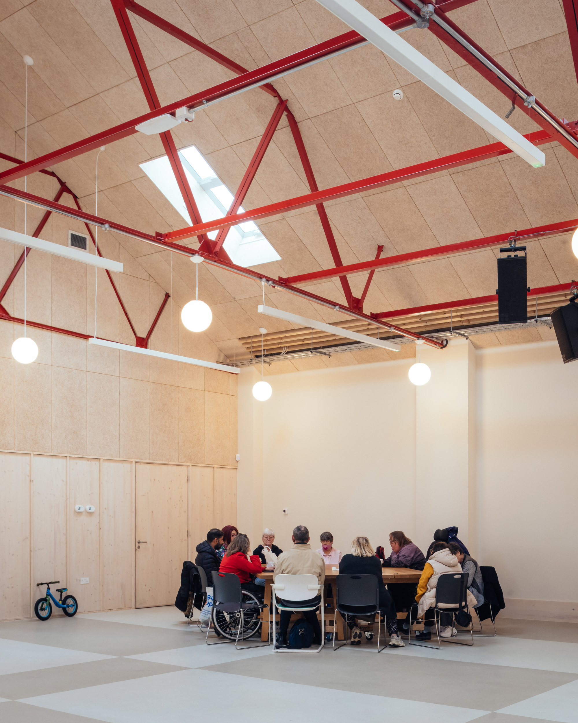 Group seated around a long table beneath red steel trusses and a rooflight, with a child's balance bike against the wall