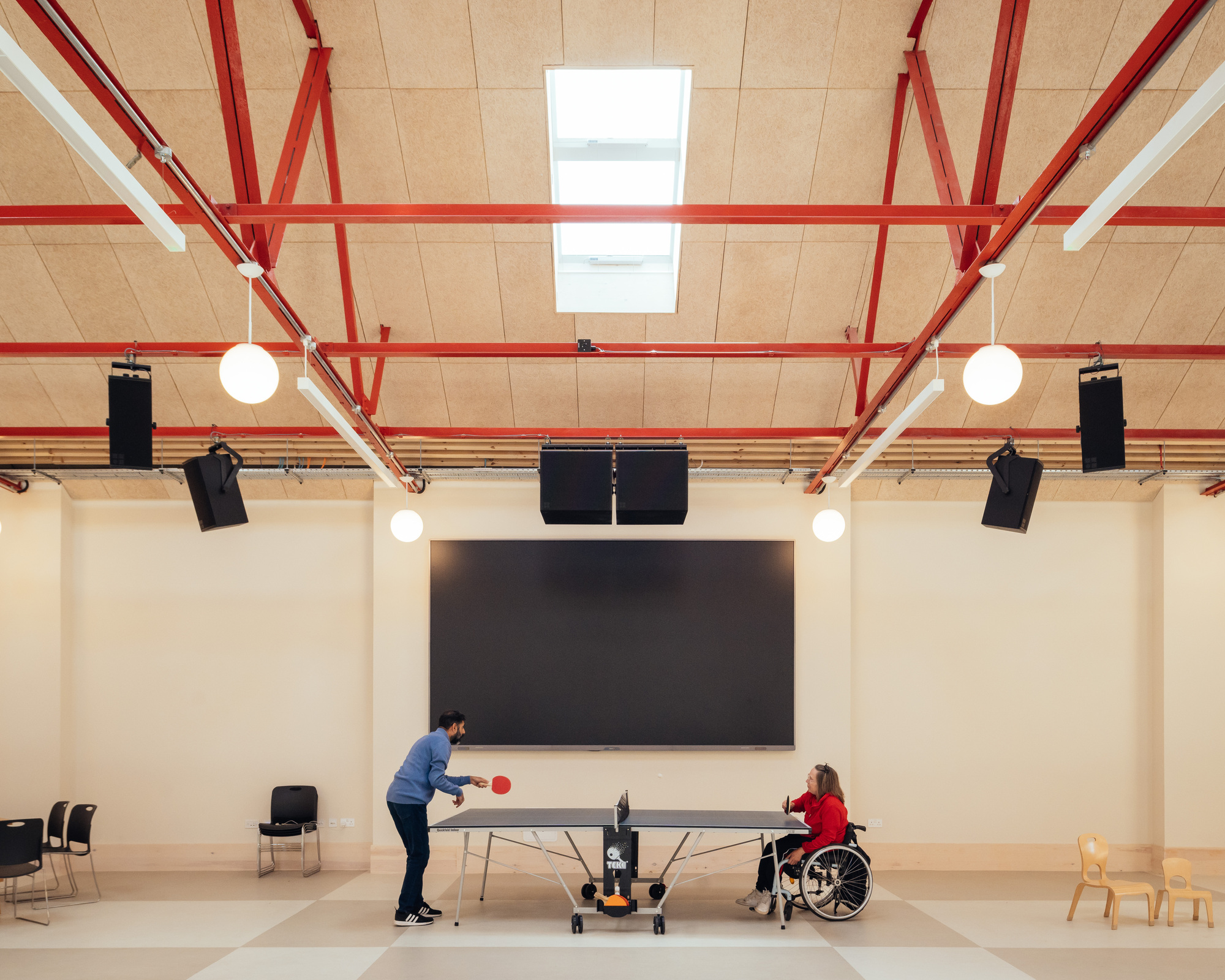 Two people playing table tennis under red trusses, with a large dark screen and hanging speakers on the wall behind